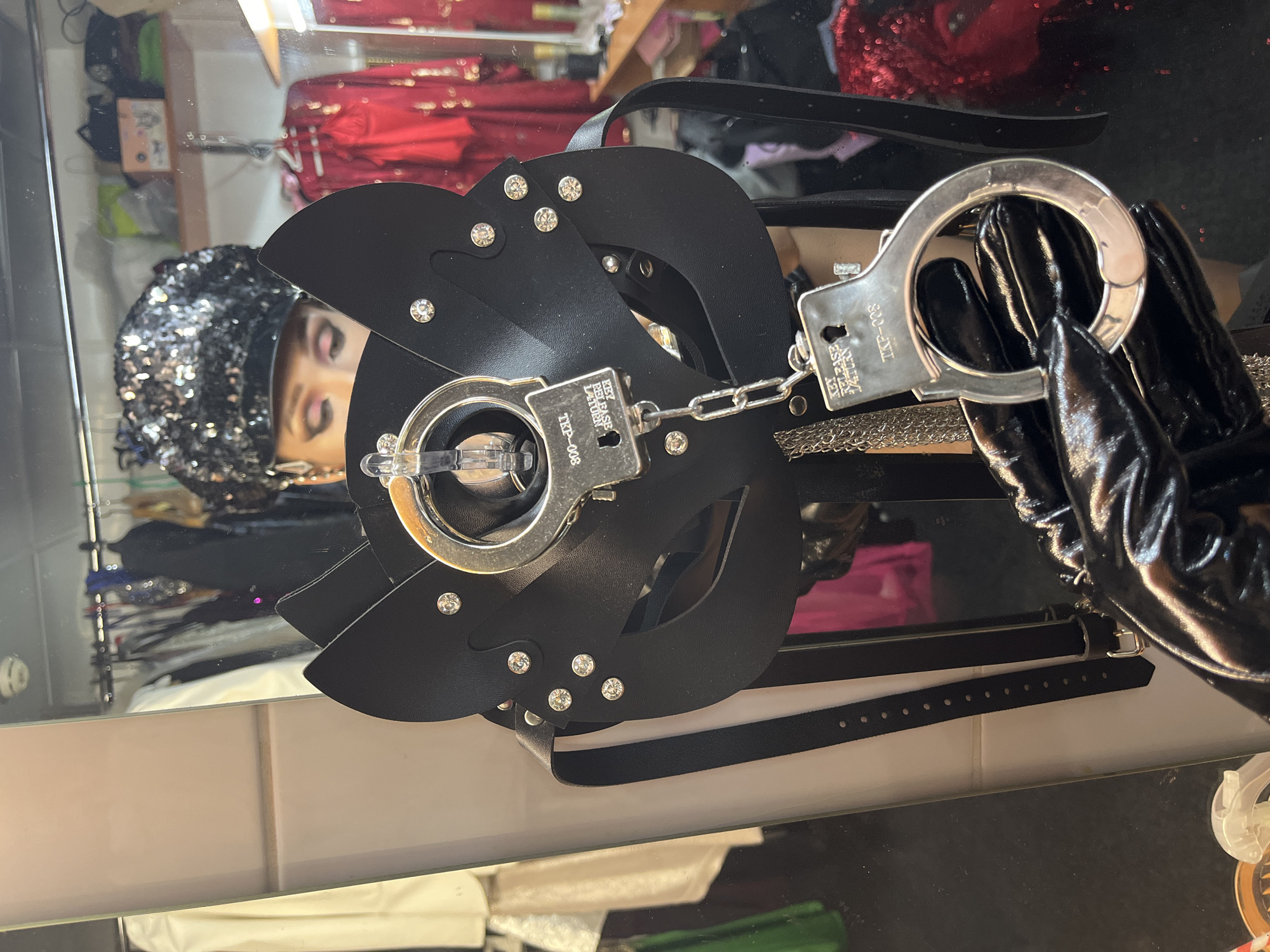 The image depicts a black leather bondage mask with metal accents and handcuffs, possibly in a fetish shop or dressing room. A black-gloved hand holds one handcuff. The background shows clothing racks and what may be a person's reflection in a mirror wearing a sequined cap. The location and the city of Kazan, Russia cannot be verified.