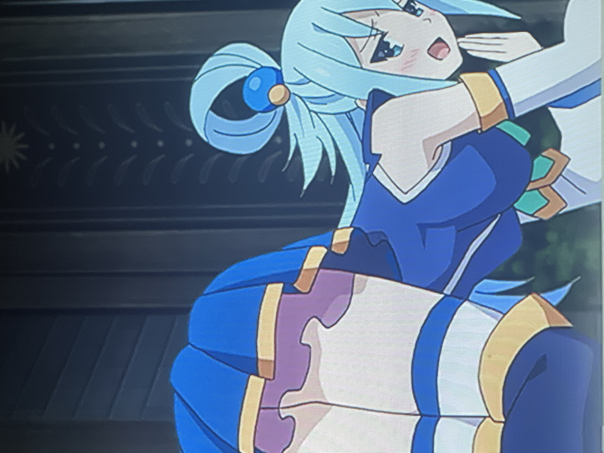 The image shows a close-up of an anime character with light blue hair and blue eyes. The character is wearing a blue outfit with gold accents. The character's mouth is open in a surprised or distressed expression, and one hand is raised to their mouth. The background is dark and appears to be a wall with some decorative elements. There is no visible indication of the city of Batumi, Georgia, or any urban environment. The scene is entirely within the frame of the animated artwork.