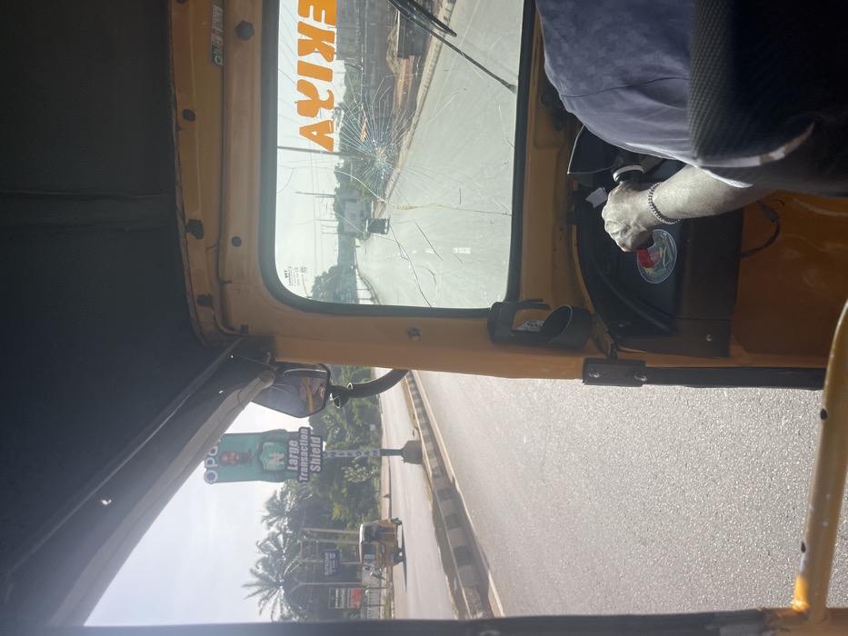 The image provides a forward-facing perspective from inside a yellow Keke Napep (tricycle taxi) on a sunny day in Barnawa, Nigeria. The driver's right arm, wearing a silver-colored bracelet, is visible on the steering handlebar. The Keke's windshield exhibits significant damage, with a prominent spiderweb fracture originating from the lower right quadrant and multiple linear cracks extending across the glass. The word "AYIKE" is visible in reversed orange lettering, appearing as "EKIYA," on the upper right interior of the windshield.
Outside the Keke, a wide, paved road with a central median is visible, featuring white dashed lane markings. Another yellow Keke Napep is observed traveling ahead in the same direction on the left side of the road. Further along the left roadside, a tall billboard displays a man's face on a green background, with the partially visible text "Opay," a large "N" symbol, and the phrase "Large Transaction Shield." Palm trees and other vegetation are visible behind the billboard. In the distance, a dark-colored vehicle is also proceeding along the road. The right side of the road reveals an undeveloped or construction area with piles of aggregate and concrete blocks. The sky above is clear.