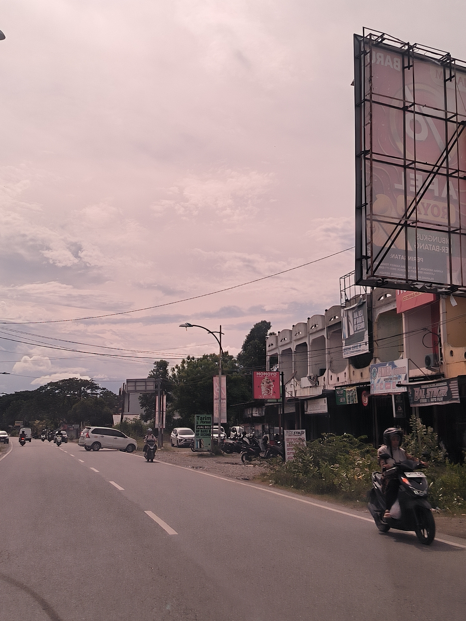 This photograph captures a bustling daytime street scene in Cot Teungoh, Indonesia, under an overcast sky filled with diffused clouds. The image is composed of a main road in the foreground and midground, flanked by commercial buildings and lush greenery.
In the immediate foreground, a paved asphalt road with clear white dashed lane markings stretches into the distance. A person wearing a helmet and a light-colored top is riding a black scooter on the right side of the road, heading away from the viewer.
The midground is filled with traffic, including several white cars, predominantly MPVs, and numerous motorcycles. Trees and dense foliage line parts of the roadside, particularly on the left. On the right side of the road, a row of commercial buildings, some featuring distinct arched upper facades, house various businesses. Overhead, a network of utility poles and power lines crisscrosses the sky, along with a streetlamp fixture.
A large, prominent billboard structure stands on the far right, displaying mirrored and largely indecipherable text, including fragmented words like "RAB" and "YOR." Additional signage on the commercial buildings is visible, including a green sign for "Tarim Ponsel" advertising "JUAL BELI HP BARU & HP SECOND" (buy and sell new and second-hand mobile phones). Other signs read "MOMOYO" on a red background, and "BAL STIKER STIKER & AUTO FILM" which appears on multiple signs, indicating a business related to stickers and car film. The overall atmosphere suggests a typical commercial area during daylight hours, with moderate traffic.