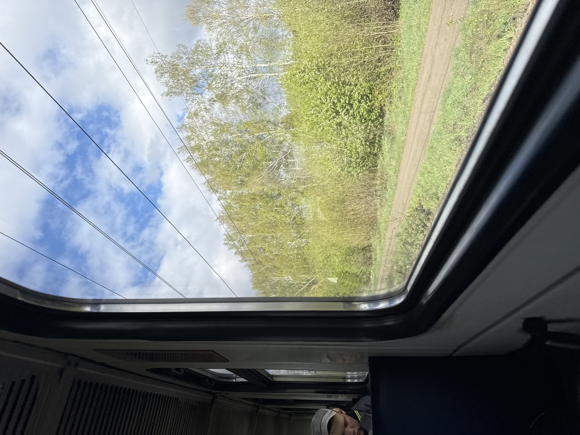 The image shows a view from inside a vehicle, possibly a bus or train, through a large window. Outside, there are trees with light green foliage and a dirt or gravel path running alongside them. The sky is visible with patches of blue and white clouds, and overhead power lines stretch across the frame. Inside the vehicle, there are interior panels and a seating area. A person's head is visible in the bottom part of the image; the person is wearing a grey hat and dark clothing. It is not possible to verify the location of the city of Mysy, Russia.