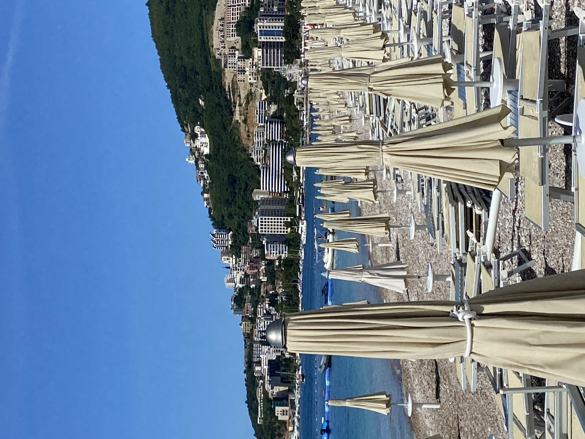 The image captures a vibrant coastal scene, likely a resort beach, under a clear blue sky. In the foreground and middle ground, a vast expanse of a pebbly beach is visible, densely lined with numerous rows of folded beige beach umbrellas and accompanying white beach chairs or loungers. The umbrellas are neatly tied, suggesting they are either closed for the day or awaiting guests. No people are visible on the beach or using the facilities.
Beyond the beach, the calm, deep blue waters of the sea stretch out, with several small boats or buoys scattered across its surface. The background is dominated by a lush, green, tree-covered hillside that rises steeply from the water's edge. Clinging to this hillside are numerous buildings, forming a coastal townscape. These structures vary in size and style, including multi-story modern hotels or apartment blocks with balconies and large windows, some featuring distinct architectural elements like black and white striped facades. The buildings appear to be predominantly white or light-colored, contrasting with the dark green of the vegetation. The sky above is a brilliant, cloudless blue, indicating clear and sunny weather, likely during the day. The scene evokes a sense of readiness for a bustling summer day, characteristic of a Mediterranean resort town like Budva, Montenegro. There is no visible text in the image.