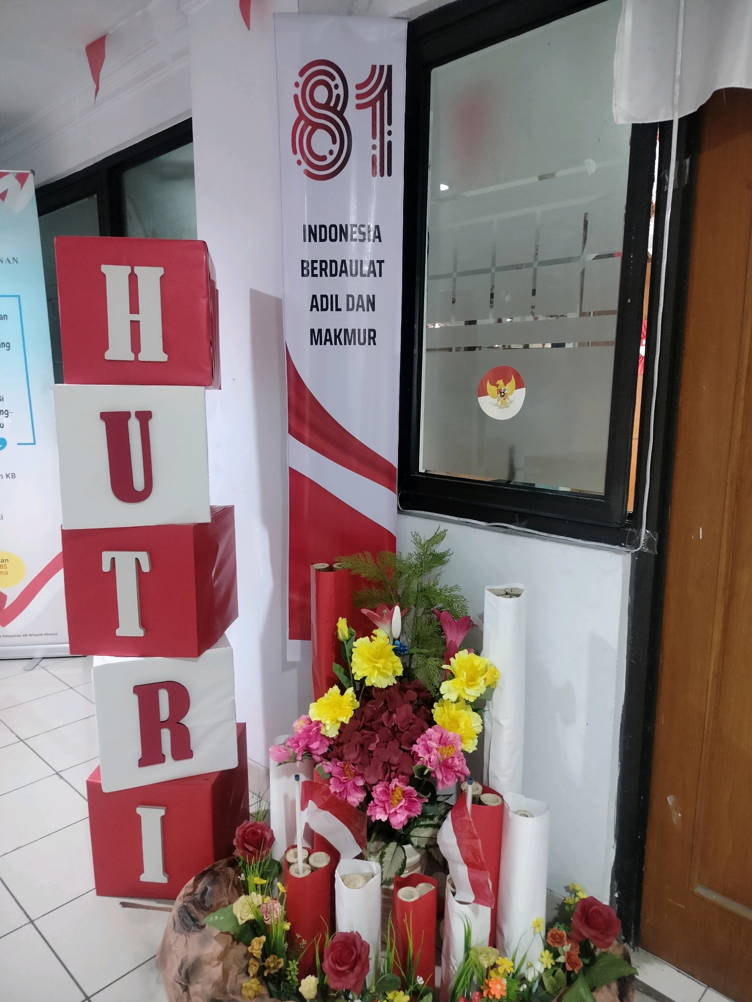The image shows a festive display in what appears to be an office or indoor public space, possibly in East Jakarta, Indonesia, given the prominent Indonesian flag colors and national emblem. In the foreground, there are stacked cubes with letters spelling "HUT RI" in white and red, a common way to celebrate Indonesia's Independence Day. Adjacent to the cubes is a floral arrangement featuring red and yellow artificial flowers, along with some red decorative cylinders and white rolls, possibly representing celebratory decorations or supplies. A banner in the background displays the number "81" in large, stylized font, along with text that appears to read "INDONESIA BERDAULAT ADIL DAN MAKMUR," which translates to "Sovereign Indonesia, Just and Prosperous." The Indonesian coat of arms, Garuda Pancasila, is visible on a frosted glass partition in the background. The overall scene suggests a celebration of a significant national event, likely related to the 81st anniversary of Indonesia's independence. The environment is indoors with tiled flooring and a wooden door visible on the right. There are no people visible in the image.