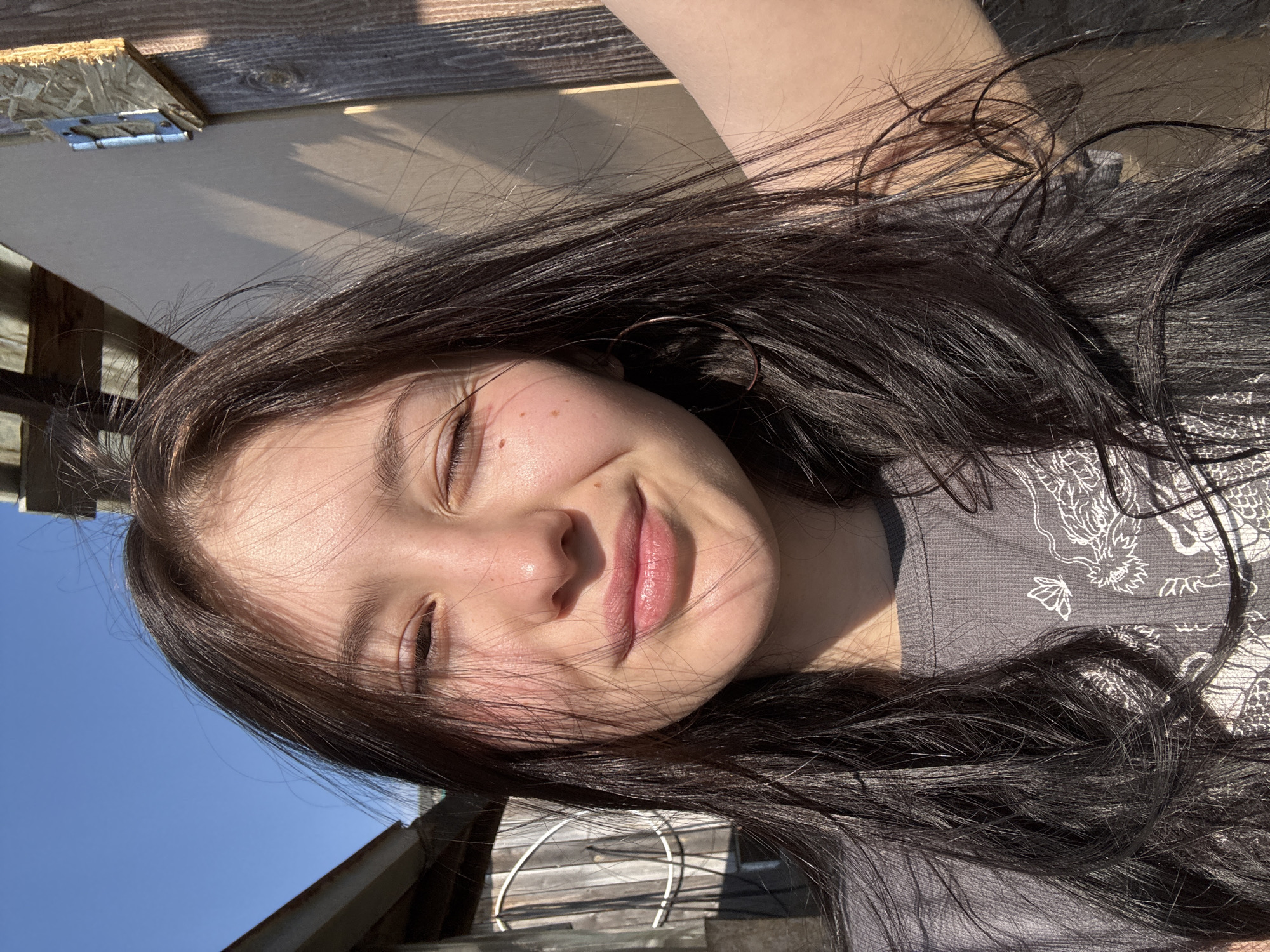 A young woman with long, dark hair is smiling with her eyes closed. She has fair skin with some freckles and is wearing a gray shirt. The background is a mix of sky, a wooden structure, and what appears to be an outdoor setting. The image does not contain sufficient information to locate the scene in the city of Фоки, Russia.