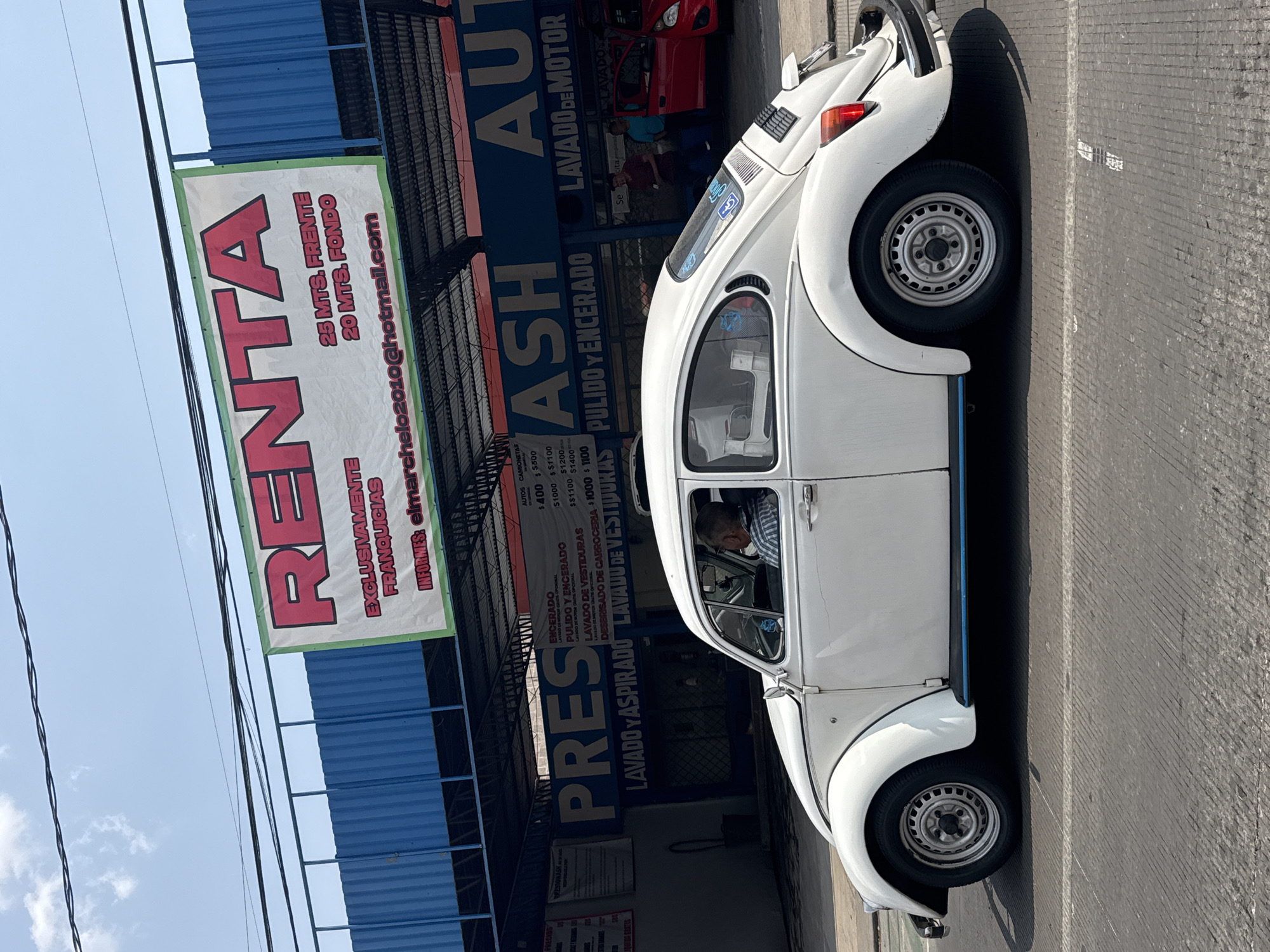 The image shows a white Volkswagen Beetle parked on a street in front of an auto detailing shop. The business signs are partially visible and indicate services such as car washing ("LAVADO") and waxing ("PULIDO Y ENCERADO"). A large banner advertises space for rent ("RENTA"). A man can be seen sitting in the driver's seat of the Beetle. The location appears to be in Puebla, Mexico.