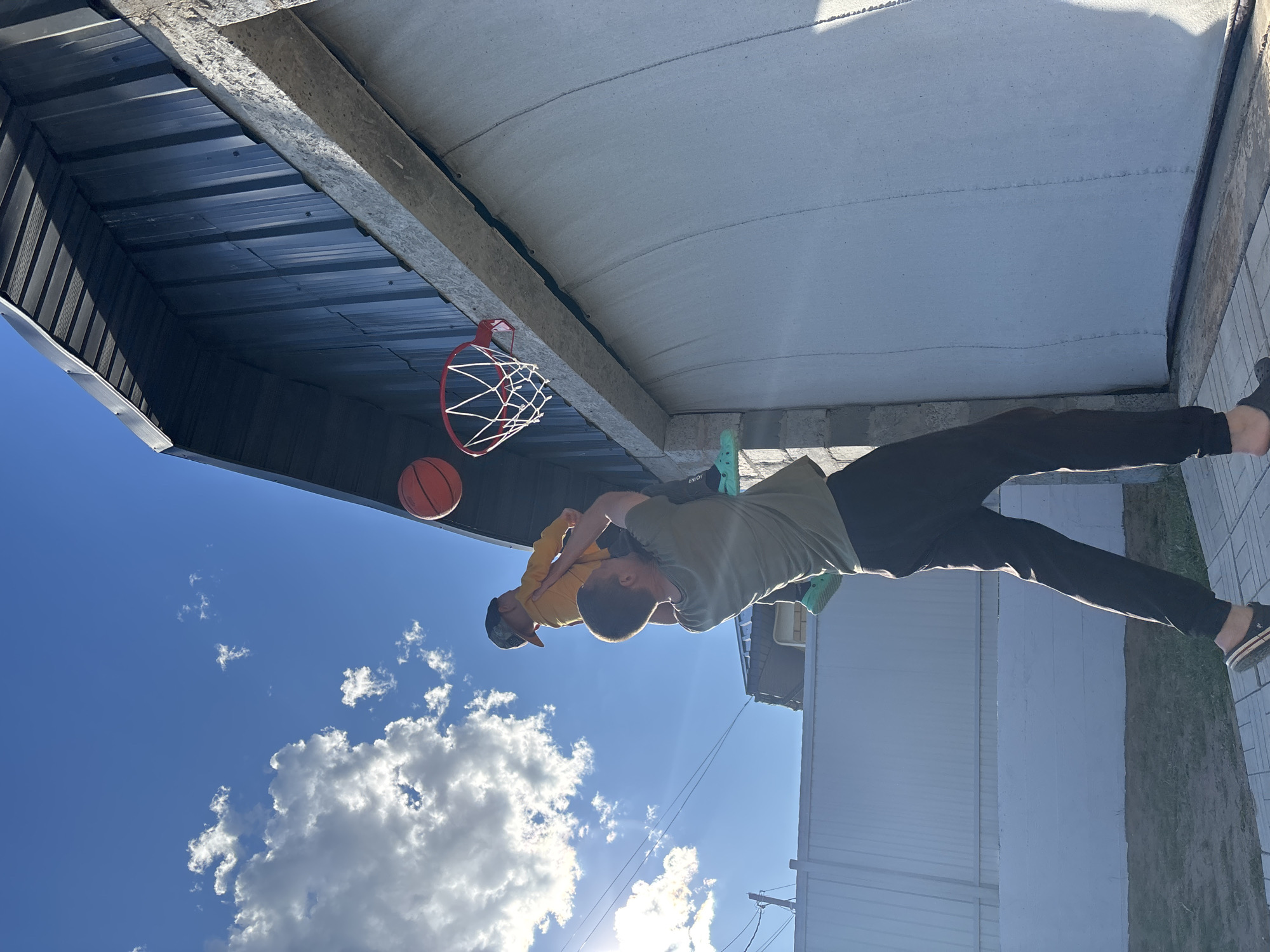 Two individuals are engaged in a physical interaction beneath what appears to be an overhang, with a basketball hoop and ball visible nearby. One person is wearing a yellow shirt and a hat, seemingly being carried or supported on the back of the other individual who is dressed in a green shirt and dark pants. The setting features a building and clear sky with clouds. The location is in the city of Chaikovsky, Russia.