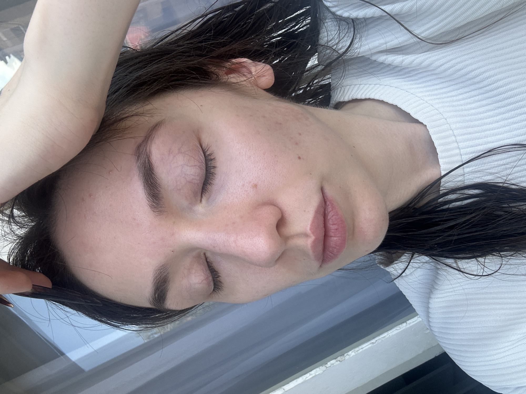 A close-up, slightly above eye-level shot shows a young woman's face with her eyes closed. She has dark, wet hair that is partially pulled back by her right hand, which is resting on the top of her head. The woman is wearing a white, ribbed top. Her skin appears fair with some freckles and minor blemishes. The background consists of a window with vertical blinds, suggesting an indoor setting. No specific location in Vracar Urban Municipality, Serbia can be determined from the image.