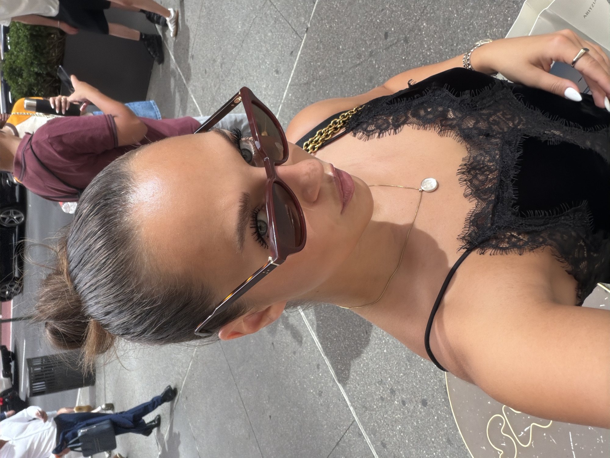 A woman is taking a selfie outdoors, with a blurry background of a city street. She is wearing sunglasses, a black lace camisole, and a gold necklace. The pavement is grey and textured. In the background, people are walking, and there are vehicles and a trash can visible. The scene appears to be a public sidewalk in New York City.