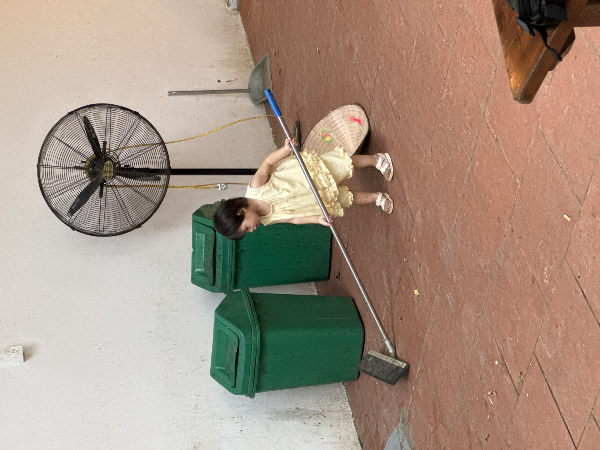 A young child in a light yellow dress and sandals is sweeping a reddish-brown tiled outdoor area with a long-handled broom. Behind the child are two large green trash cans and a large industrial fan mounted on a stand. The wall behind the objects is a rough, off-white texture. A woven basket or hat is lying on the ground near the child. The scene appears to be in an outdoor, possibly covered, area with a concrete floor. The general ambiance suggests a casual, everyday setting, perhaps in a residential or communal space.