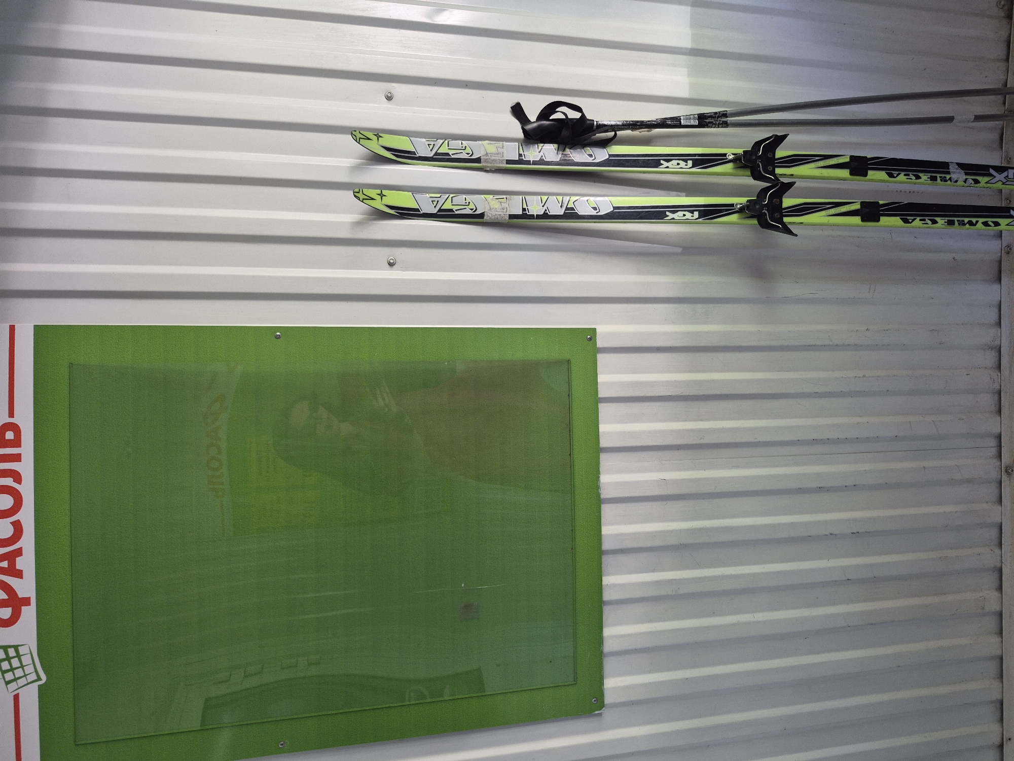 The image shows a wall with a green board on the left and a pair of skis with poles mounted above it. The wall appears to be made of corrugated metal sheets. The skis are predominantly yellow and black with "OMEGA" printed on them. The location is Unknown, Unknown.
