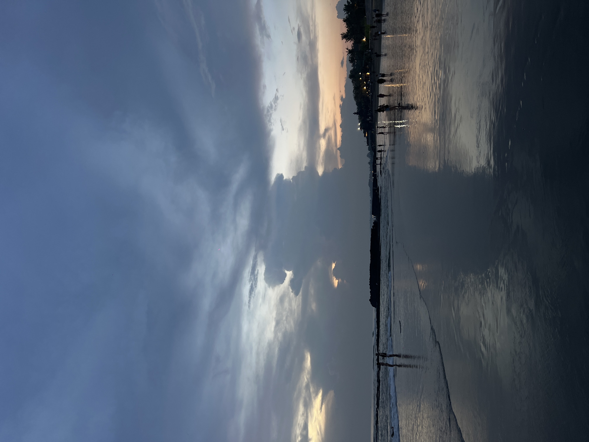 The image shows a beach scene at sunset in Canggu, Indonesia. The sky is filled with clouds, varying in color from dark blue to orange. The ocean reflects the colors of the sky. Silhouettes of people can be seen walking along the beach. There are buildings and trees in the distance.