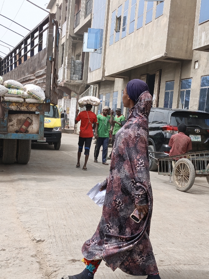 The image captures an urban street scene in Fagge C, Nigeria, during daylight. In the foreground, a woman walks from right to left, observed from her left side. She wears a long, patterned dress in maroon, black, and white, topped with a dark blue hijab. Her left hand holds multiple white papers, while her right hand grips a light-colored smartphone. Her left lower leg is encased in a dark cast or brace, adorned with colorful fabric elements around the ankle.
In the middle ground, a man in a red t-shirt and dark shorts walks away from the viewer, balancing a large white sack on his head. To his right and slightly behind him, several other men are visible; at least three wear green t-shirts, with some wearing white caps.
To the far left, the rear section of a large, blue utility truck is present, its bed loaded with multiple white sacks of goods. A yellow compact utility vehicle is partially visible behind the truck. On the right side of the frame, a black SUV with a "LAGOS TYU 964HY" license plate is parked. Behind the SUV, a man in a red and black patterned shirt sits on a wooden handcart featuring large, spoked wheels.
The background consists of multi-story buildings; some display modern blue-tinted glass windows, while others exhibit exposed concrete and rebar, suggesting ongoing construction or various states of repair. The ground is a light-colored, paved surface. A metal structure providing overhead cover is partially visible in the upper left.