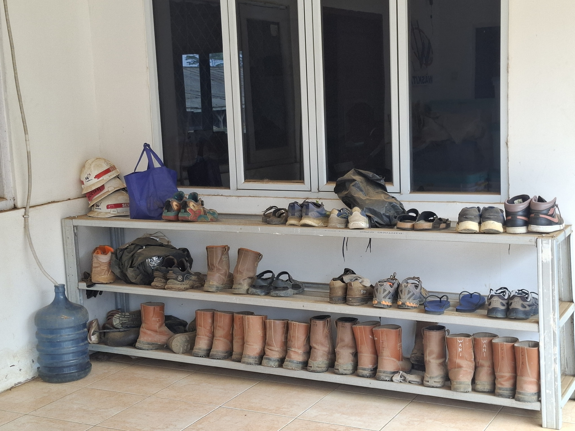 This image depicts an outdoor storage area likely situated in Ciheulang Tonggoh, Indonesia, characterized by a tiled floor and a white wall with a large window featuring multiple panes. A three-tiered metal shelving unit is prominently displayed, laden with various footwear and accessories.
On the top shelf, several pairs of boots, including work boots and what appear to be hiking boots, are neatly arranged. Next to them are casual shoes, sandals, and a dark plastic bag. On the left side of the top shelf, a stack of white hard hats with red and black detailing are visible, along with a blue tote bag.
The middle shelf holds an assortment of footwear, including boots, sandals, and athletic shoes. Some items are dusty, suggesting their use in an outdoor or work environment. A large blue water jug with a tube extending from its opening is positioned on the floor to the left of the shelving unit.
The bottom shelf is densely packed with an array of work boots, primarily in shades of brown and orange. Many of these boots are scuffed and show signs of wear.
The overall scene suggests a practical storage solution for protective gear and footwear, commonly found in residential or work settings in regions like Ciheulang Tonggoh. The presence of work boots and hard hats indicates a potential connection to manual labor or construction. The environment is clean and organized, with the shelving unit serving as a central focal point for the items.