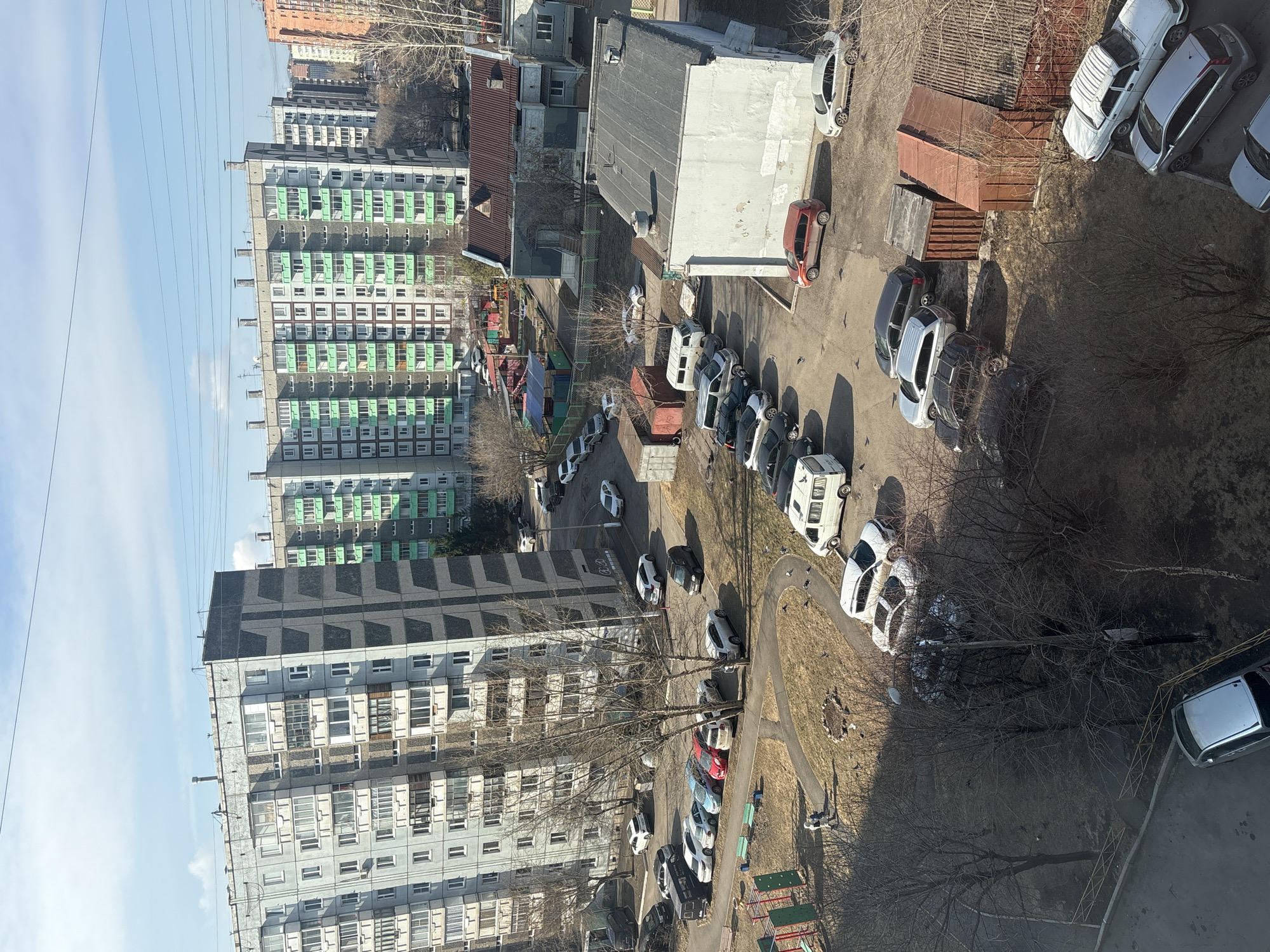 The image depicts a densely populated residential area in Krasnoyarsk, Russia, characterized by Soviet-era apartment blocks. The buildings are primarily constructed of concrete and feature a repetitive pattern of windows and balconies.
A large parking lot occupies the center of the image, filled with numerous cars of varying makes and models. Several large metal containers are scattered around the parking area, potentially used for waste disposal or storage.
A small playground is visible among the apartment buildings, containing swings, slides, and other play structures. A few bare trees are scattered around the scene. The overall impression is one of a typical urban landscape in a post-Soviet city. The lighting suggests it is midday.