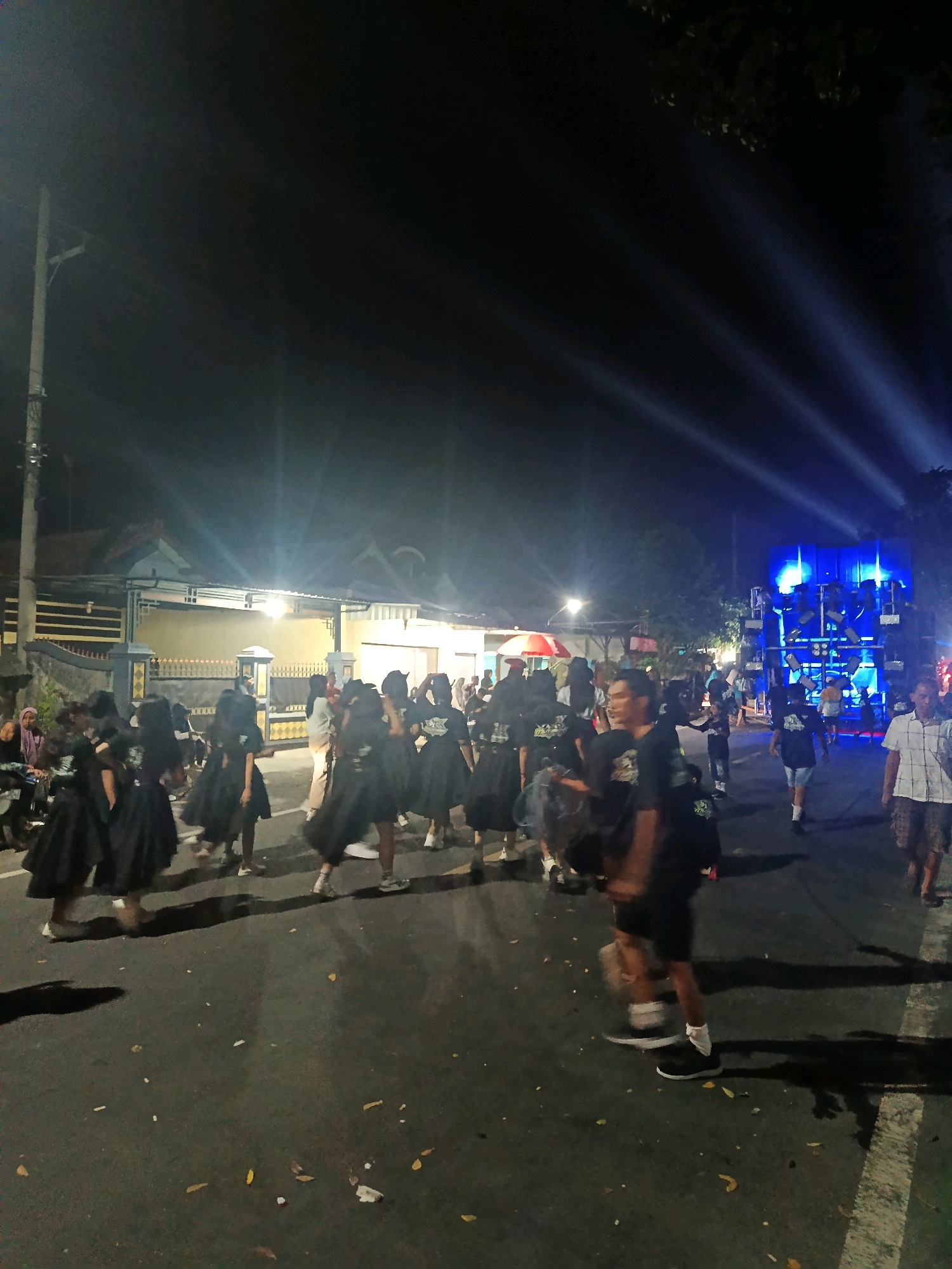 The image depicts an outdoor nighttime scene in Pasirian, Indonesia, likely on a street. A group of individuals, predominantly young people dressed in black, are visible, some in black dresses and others in black t-shirts. They appear to be participating in a procession or performance, possibly related to a cultural event or festival. In the background, a large blue sound system or stage is illuminated by blue lights, with beams of light projecting upwards into the dark sky. A few buildings with lights are also present along the street. Some bystanders, dressed in casual attire, are also present, observing the activities. The ground is asphalt, with scattered leaves and debris. The overall atmosphere suggests a lively, public gathering at night.
