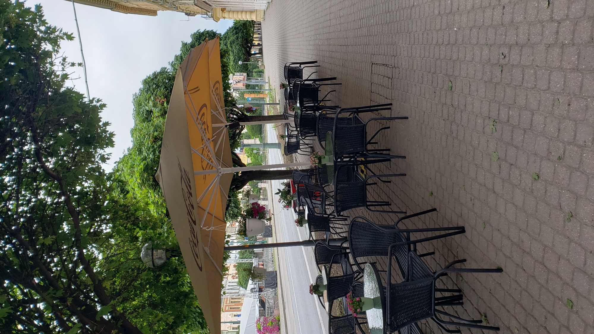 The image depicts an outdoor seating area of a cafe or restaurant located on a sidewalk in Prešov, Slovakia. The scene is characterized by numerous black, woven-style chairs and small, round glass-topped tables arranged under a large, tan umbrella advertising Coca-Cola. Hanging flower baskets with pink and white flowers are visible, adding a touch of color to the area. The sidewalk is paved with grey brickwork, and fallen leaves are scattered across it. Adjacent to the sidewalk is a street with traffic, and across the street, there are buildings and a large white tent structure, possibly for an event. Trees with lush green foliage line the sidewalk and the street, providing shade. The sky is overcast. There are no people visible in the image. The setting appears to be a public area in a European city, consistent with Prešov.