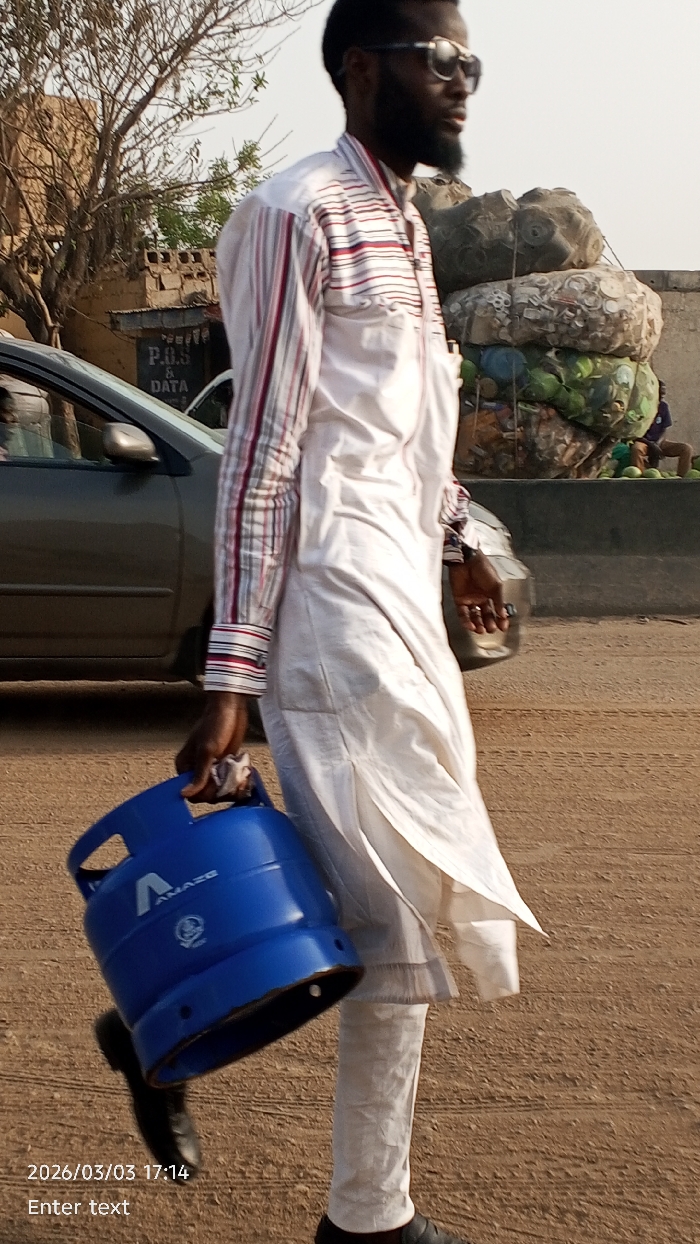 An adult male with dark skin and a beard, wearing sunglasses, a striped long-sleeved shirt over a white tunic, and white trousers, walks from left to right on an unpaved, light brown-orange dirt road in Tudun Wada, Nigeria. He carries a blue "AMAZE" brand gas cylinder in his left hand. In the background on the left, a dark-colored sedan is partially visible, behind which stands a building with a "P.O.S & DATA" sign and a bare tree. To the right, a tall stack of various bundled items, possibly containing recyclables or produce, is situated in front of a light-colored wall. Indistinct figures are present at the base of the stacked items. The scene is illuminated by natural light, consistent with the timestamp 2026/03/03 17:14.