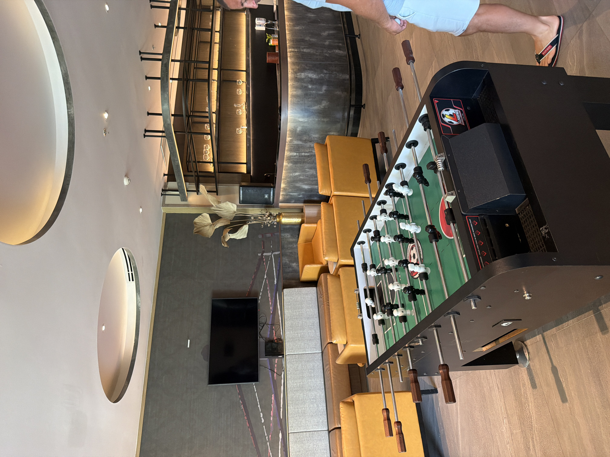 The image depicts an indoor recreational area, possibly a lounge or game room, in Bang Muang, Thailand. In the foreground, a black foosball table with green turf and white and black player figures is prominently displayed. The table has wooden handles and metal rods. To the right of the foosball table, a person wearing light blue shorts and flip-flops is visible from the waist down, standing and facing the table.
The background features a modern lounge setting with several yellow upholstered modular sofas and chairs arranged around the foosball table. Behind the seating area, a large flat-screen television is mounted on a dark grey textured wall. Further back, a stylish bar area with a dark countertop and illuminated shelves displaying glassware is visible. A decorative plant with large, dried leaves stands near the bar. The ceiling has an interesting architectural design with a circular recessed lighting feature and an air vent. The flooring is tiled with a light grey, wood-grain pattern. The overall impression is a contemporary and comfortable space designed for relaxation and entertainment.