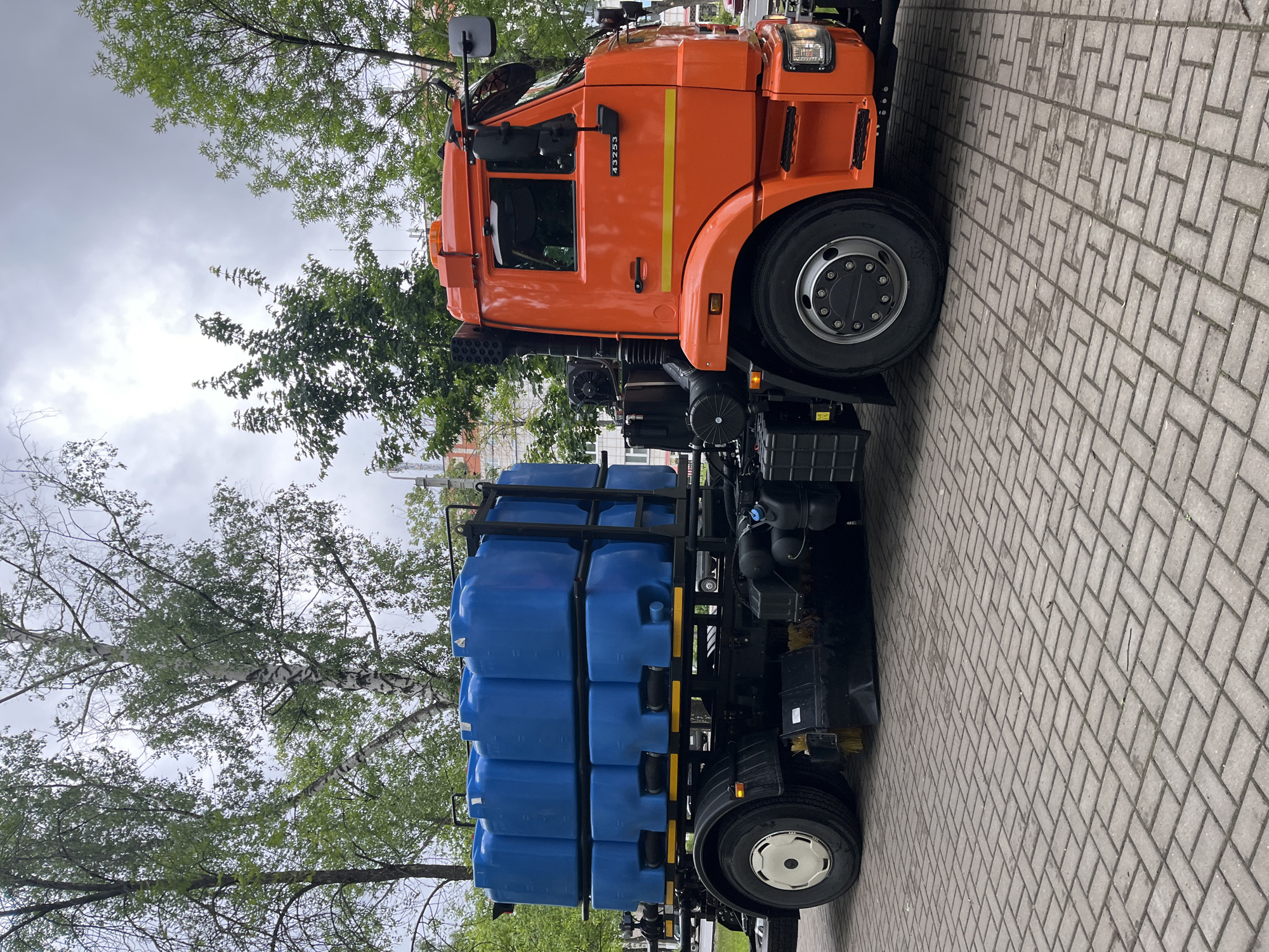 A large, orange heavy-duty truck, identified as a KAMAZ vehicle, is parked on a paved surface in an outdoor setting, likely in Chaikovsky, Russia. The truck features a prominent orange cab with a yellow vertical stripe on its side, bearing the number "43253". The rear section of the truck is equipped with a specialized black metal frame, which holds multiple blue rectangular plastic tanks or containers, stacked in rows and columns. Black hoses or pipes are connected to some of these tanks, indicating the vehicle is designed for liquid transport or distribution. Additional equipment, including a black storage box and what appear to be brush attachments, are visible near the rear wheels, suggesting it may be a municipal service or cleaning vehicle.
The truck is stationary on grey herringbone-patterned pavers. Surrounding the vehicle are deciduous trees with lush green leaves, implying a spring or summer season. The sky is overcast and cloudy, providing soft, diffused daylight without strong shadows. No people are visible, and no specific activity is taking place beyond the truck being parked. The scene conveys a functional and operational environment for a specialized industrial vehicle.