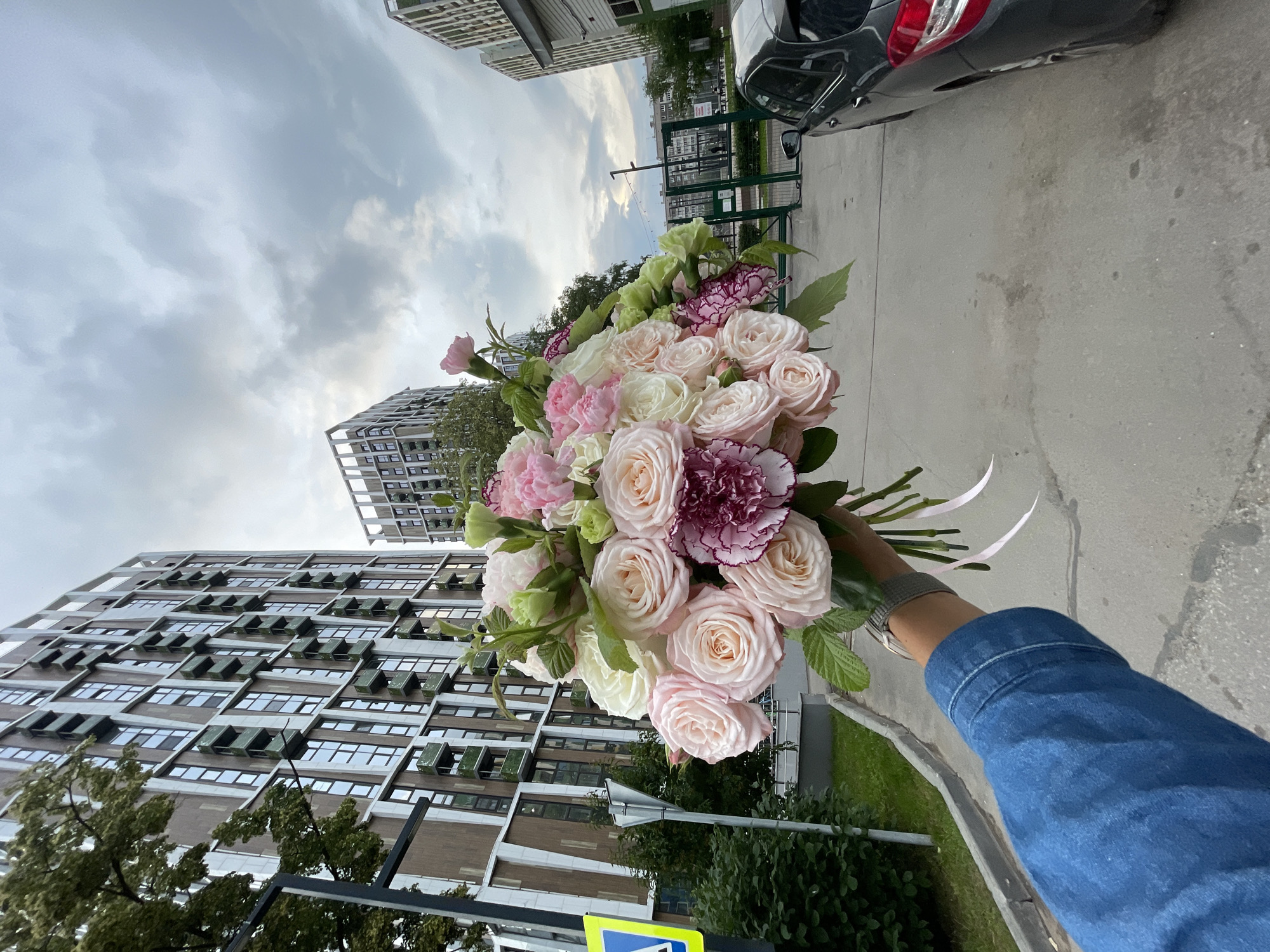 The image features a person holding a bouquet of flowers. The bouquet is composed of roses in various shades of pink and white, with some green foliage and dark pink carnations interspersed. The person's arm is visible, wearing a blue denim jacket, and a watch on their wrist. The background depicts an urban environment with modern, tall buildings and a cloudy sky. A parked car is also visible on the right side of the frame. The perspective is from the person's point of view, looking upwards towards the buildings and sky, with the bouquet held in the foreground. The lighting suggests it is daytime, possibly overcast due to the cloudy sky. There is no visible text in the image.
