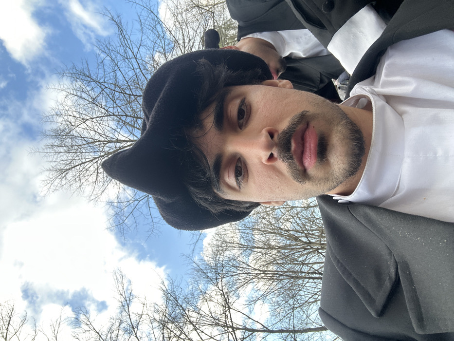 The image depicts an outdoor scene in Guimarães, Portugal, on a day with a partly cloudy blue sky and numerous leafless deciduous trees. The primary subject is an adult male, likely young, with dark hair, a prominent dark mustache, and a short beard. He is looking directly forward, possibly at the camera. He wears a black, felt-like hat with a distinctively folded or peaked crown, a black overcoat or jacket with lapels, and a white, high-collared shirt. Partially visible behind and to the right of the primary subject is another individual, also appearing male, wearing similar dark attire including a black coat and a white shirt, with a dark hat visible on their head. Both individuals are positioned within a natural environment characterized by the bare trees, suggesting a winter or early spring setting.