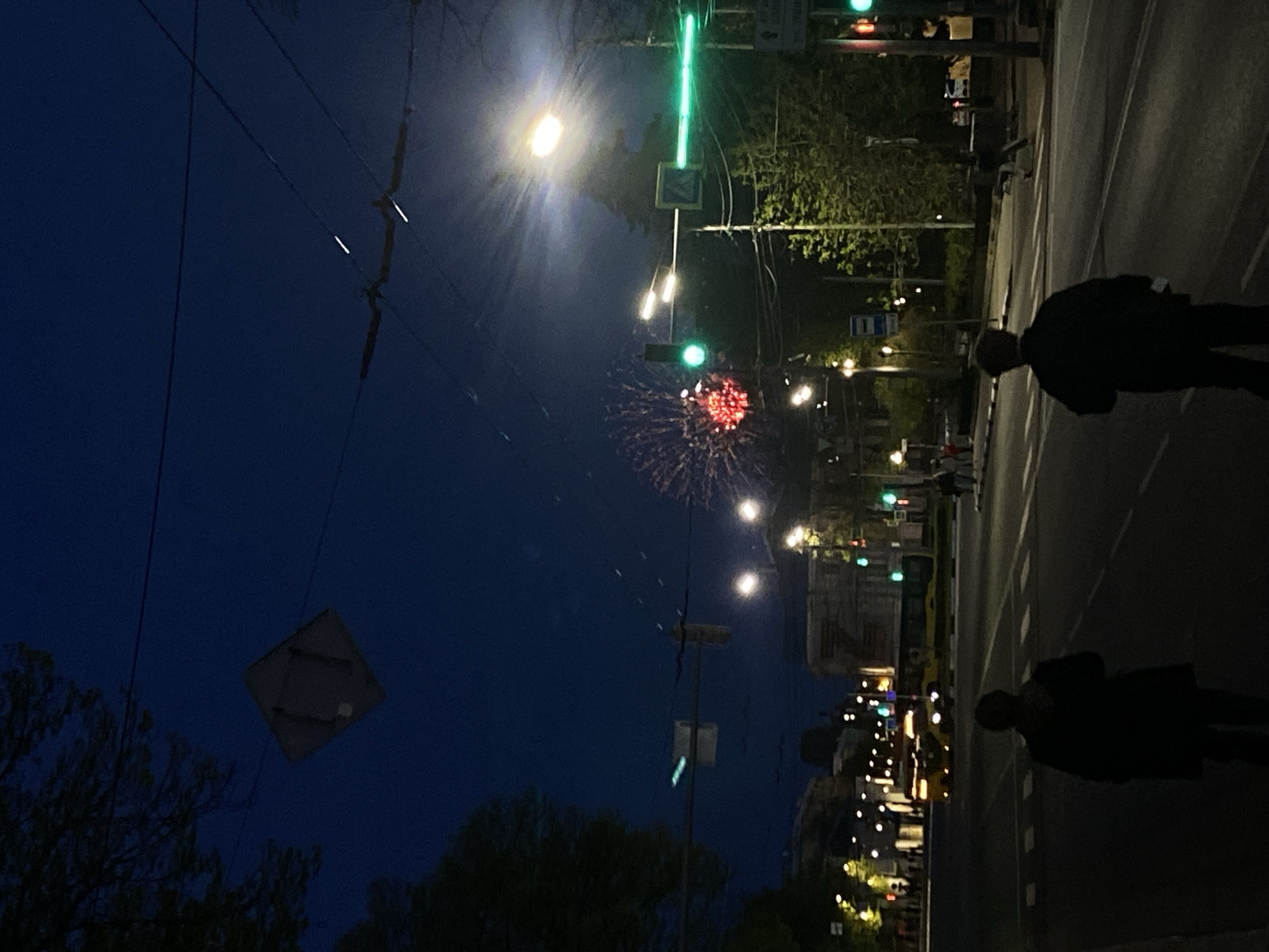 Captured at night in Шебунино, Russia, the image presents a street scene with fireworks in the sky. A pedestrian is visible on the sidewalk, and a city street with streetlights, traffic lights, and pedestrian crossing signs illuminates the scene. Buildings and trees line the street, creating a bustling urban atmosphere. Overhead wires are visible against the dark blue sky.