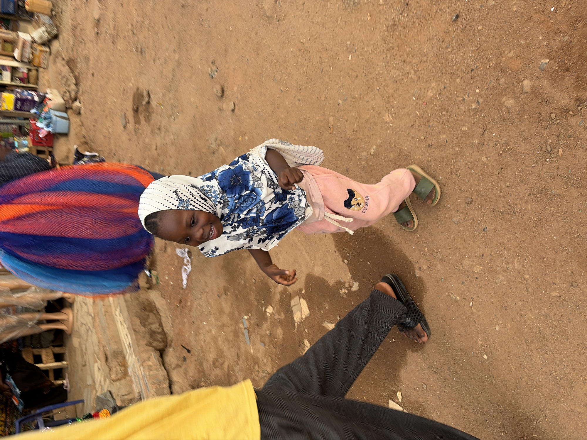 The image shows a child wearing a head covering, a blue and white floral shirt, and pink pants. They are smiling and walking on a dirt surface. In the foreground, a person's leg is visible, wearing gray pants and black sandals. In the background, there are buildings and goods displayed. The ground is dirt, and the scene appears to be in Jos, Nigeria, based on the context.