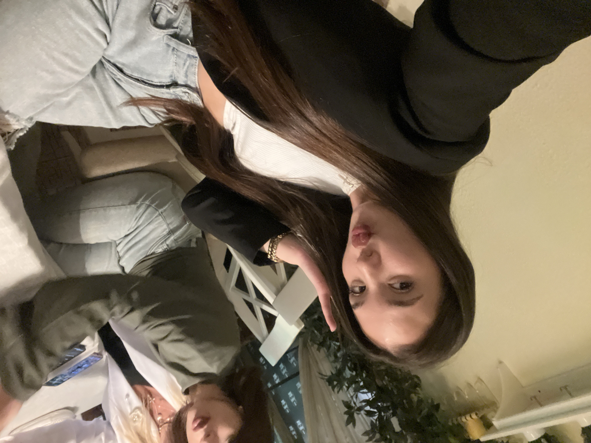 The image depicts two women in an indoor setting. One woman is wearing light-colored jeans, a white top, and a black blazer. She has long, dark hair and is sticking out her tongue. She is wearing a gold bracelet. The other woman is wearing a green jacket and a white top. A table is visible with phones and other objects on it. There is a white chair visible behind the first woman. The environment includes a light-colored wall and decorative plants. The location appears to be a restaurant or a similar establishment. There is no information in the image to confirm the location in Chaikovsky, Russia.