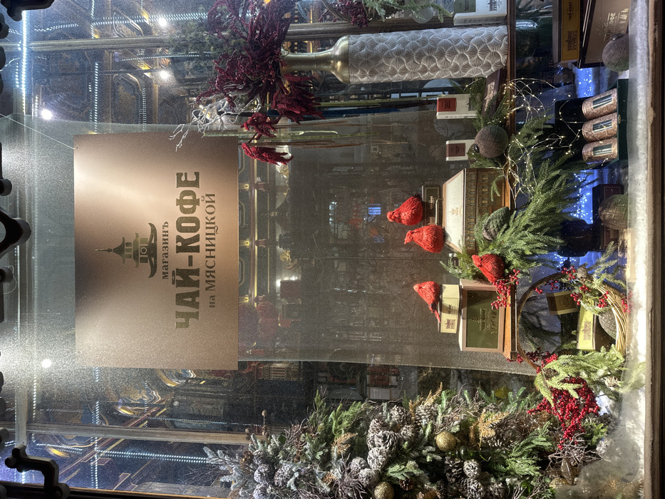 A shop window display features a sign in Russian advertising "Tea-Coffee" with an oriental-style pagoda logo. The display is decorated for the holidays with pine branches, pine cones dusted with snow, gold ornaments, and red berries. Several small red bird figures are perched on stacked boxes. A long, patterned vase and decorative balls also adorn the display. The interior of the shop is visible through the glass.