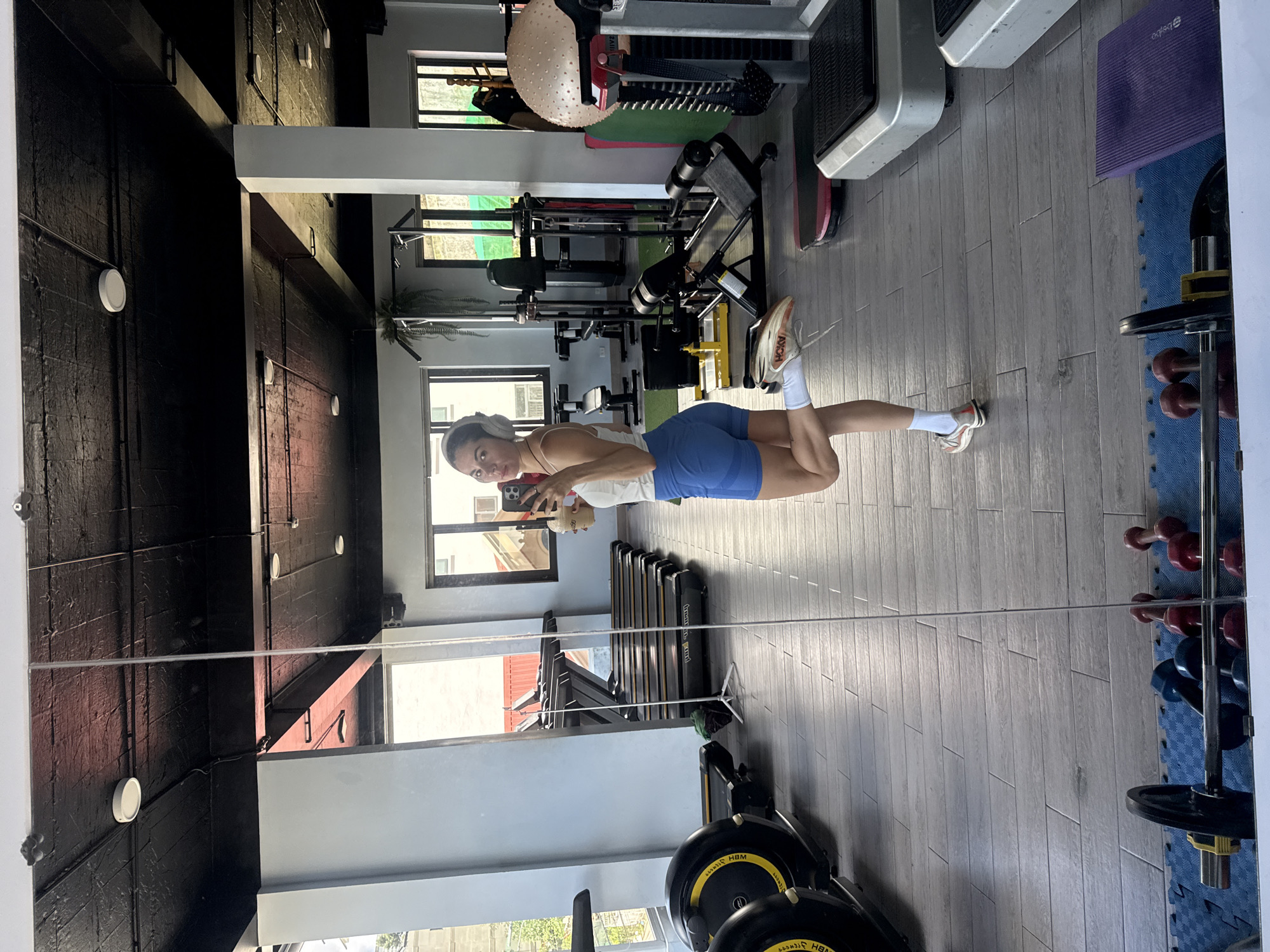 In Da Lat, Vietnam, a woman stands in a gym, posing for a selfie in a full-length mirror. She is wearing a white tank top, blue athletic shorts, white socks, and athletic shoes. She has headphones on and is holding a takeaway coffee cup in her right hand. Her left leg is bent and extended backward. The gym is equipped with various exercise machines, including treadmills, elliptical machines, weight machines, and a stability ball. The flooring is light-colored wood planks, and the ceiling is painted black with exposed pipes and light fixtures. Dumbbells and a barbell are visible on a blue mat in the foreground, reflected in the mirror. The overall scene depicts a typical gym environment with a focus on fitness.