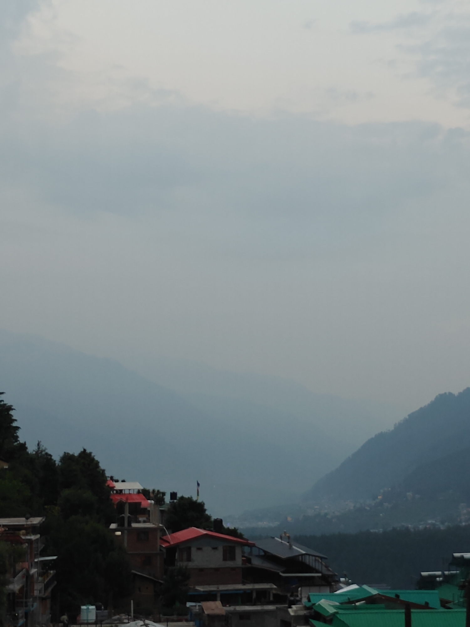 A wide shot captures a serene mountain landscape with a village nestled on its slopes. The sky is overcast with soft, diffused light, suggesting it could be early morning or late afternoon. The air appears hazy, adding a sense of depth and atmospheric perspective to the scene.
The foreground is dominated by a cluster of houses with varied roof designs, some featuring vibrant red, while others are topped with green. The architecture suggests a traditional mountain village. Dense green foliage, likely trees, flanks the structures, further emphasizing the natural setting.
In the background, layers of mountains recede into the distance, their forms softened by the haze and distance. A small town or settlement can be faintly seen in a valley or on a further slope, indicated by scattered lights. A flag flies on one of the buildings in the village, adding a touch of local detail.
The overall impression is one of peaceful coexistence between human habitation and the grandeur of nature. There are no people visible, and the scene appears still, devoid of any specific activity. The time of day is not precisely clear due to the overcast conditions, but the soft light implies a period of reduced sunlight. The location cues, such as the style of the buildings and the mountainous terrain, combined with the provided location context of Vashisht, India, firmly place the scene in a Himalayan region. There is no visible text in the image.