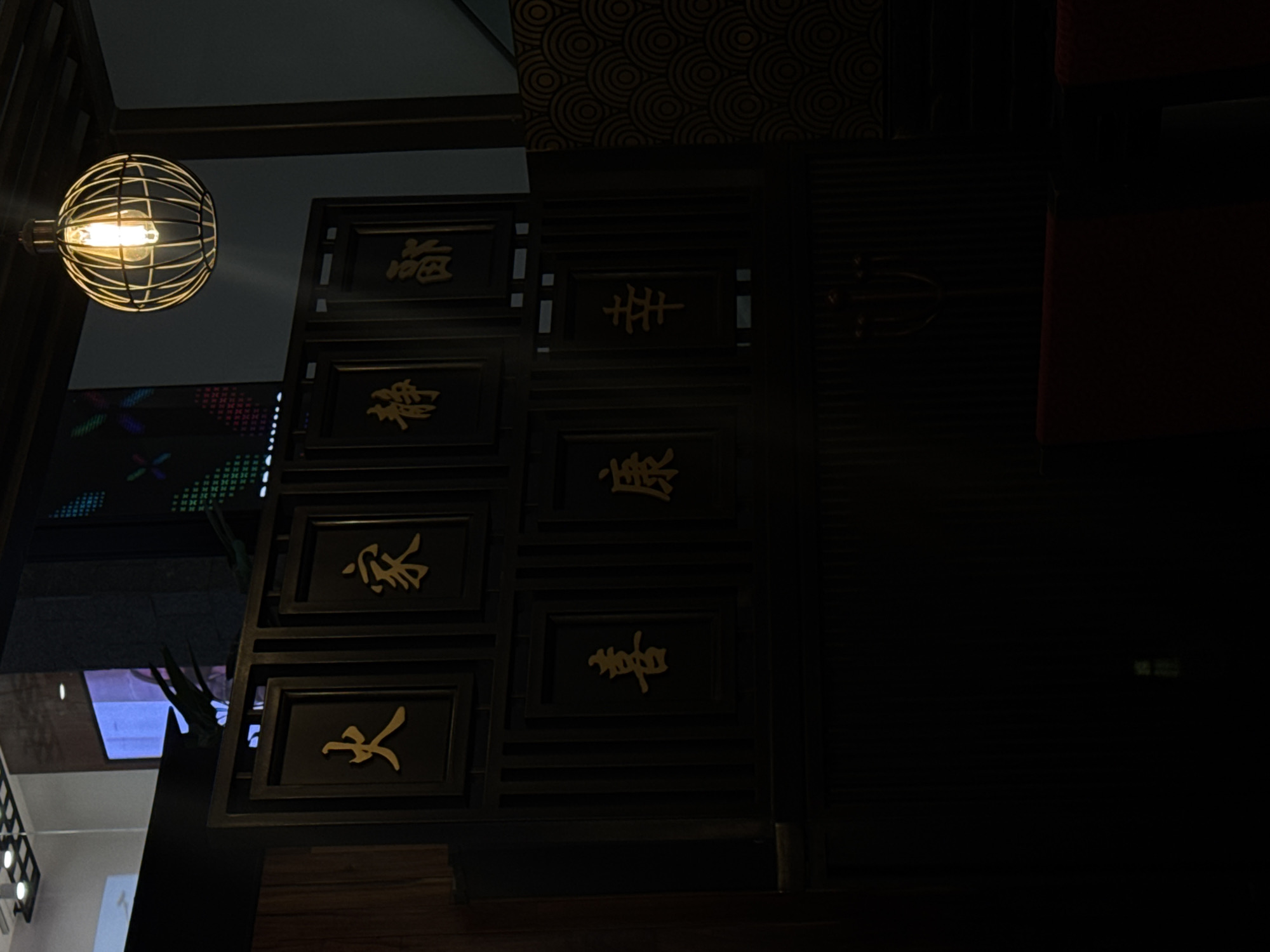 The image depicts the interior of an East Asian-themed establishment, possibly a restaurant, located in Puebla, Mexico. The scene is dimly lit, focusing on a dark wooden partition or wall panel that serves as a decorative feature. This panel features seven golden Chinese characters set within individual rectangular frames, arranged in two rows. The top row, from left to right, displays the characters 火 (huǒ - fire), 家 (jiā - home/family), 靜 (jìng - quiet/calm), and 鄙 (bǐ - vulgar/rustic/humble). The bottom row, from left to right, displays 喜 (xǐ - joy/happiness), 康 (kāng - healthy/peaceful), and 幸 (xìng - good fortune/luck). Above the panel, to the right, a spherical pendant light with a metal cage design encases a glowing Edison-style filament bulb, casting a warm light. To the right of the wooden partition, a dark wall section is visible, adorned with a repeating circular or wave-like pattern. Reflections in an overhead glass surface reveal parts of a modern interior, including bright overhead lights and a colorful digital display screen with a pixelated pattern. The lower portion of the image shows the top edge of a red upholstered piece of furniture, likely a chair or booth, slightly out of focus. No individuals are present in the scene.