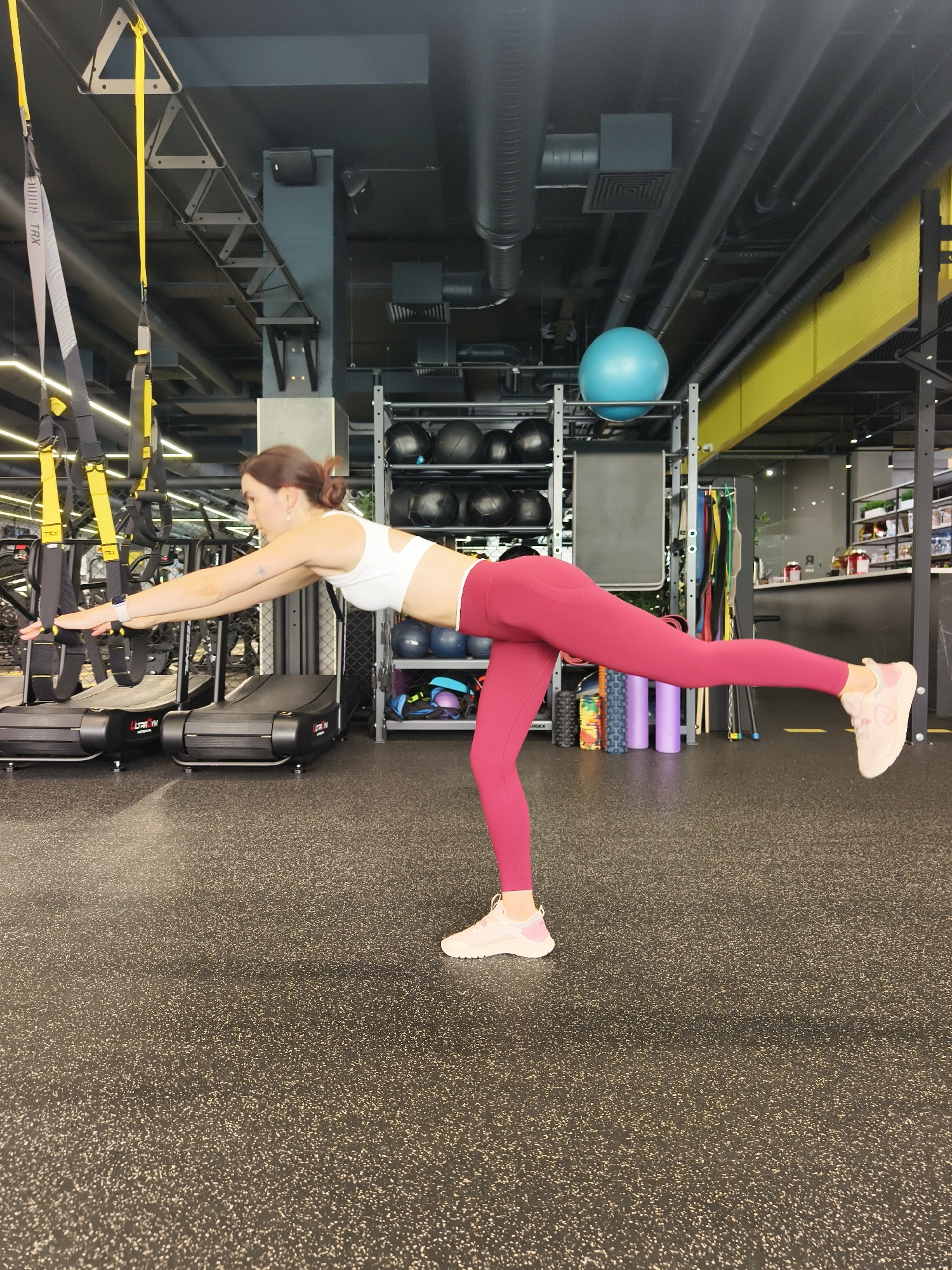 A woman is performing a single-leg balance exercise in a gym. She is wearing a white sports bra and maroon leggings. Her left leg is extended behind her, and her arms are outstretched forward. The gym has a dark, speckled floor, treadmills in the background, and various fitness equipment including a blue exercise ball, medicine balls, and yoga mats. The scene is located in a gym in Tyumen, Russia.