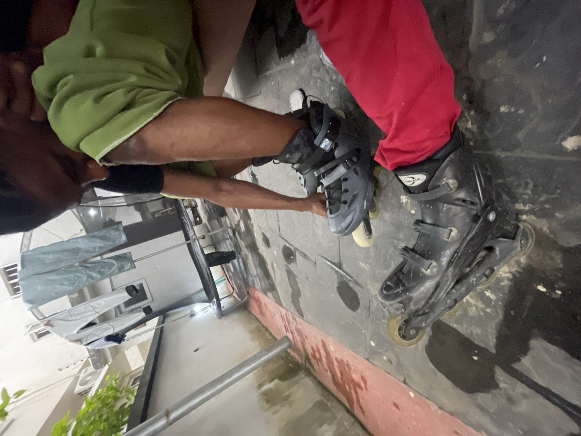 An outdoor, low-angle shot depicts two individuals in Ado, Nigeria, preparing to rollerblade. The ground is paved with dark grey tiles, wet in patches with some reddish water stains visible along a narrow strip. In the foreground, one person, wearing a bright red garment and a black rollerblade with yellow wheels, is partially visible. The other individual, positioned behind and to the right, is wearing a green hooded garment and dark shorts. This person is leaning over and adjusting their grey rollerblade, which has black accents and a white heel strap. Behind them, a clothesline is strung with several items of clothing, including light blue trousers and white garments. An outdoor trampoline with a black net is visible in the middle ground. The background shows the side of a building with windows and an air conditioning unit. The lighting suggests it is evening or dusk.