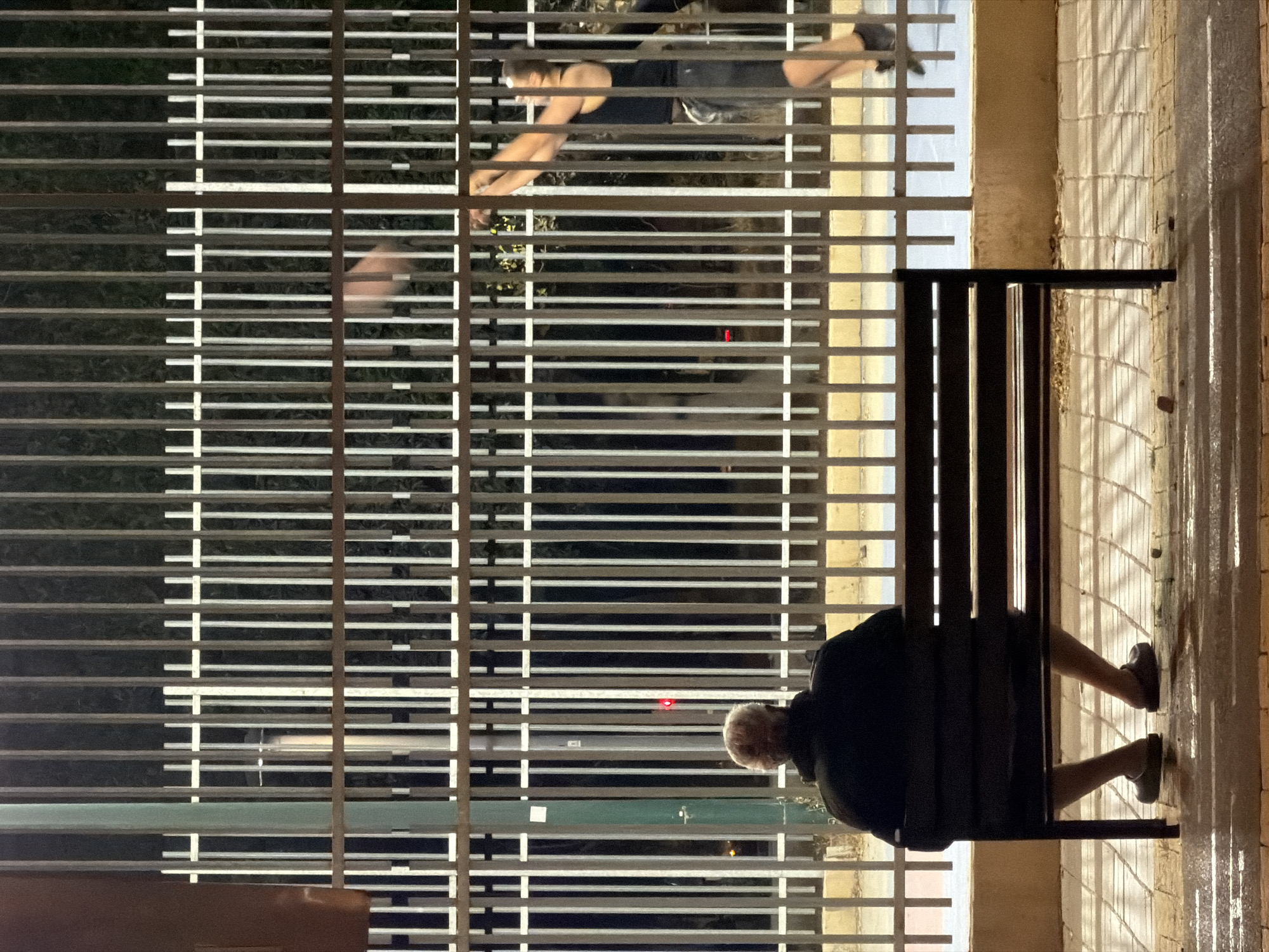 The image shows a night scene in Tel-Aviv, Israel. An elderly person sits on a dark bench. The bench is positioned next to a metal barrier of horizontal bars and vertical supports. On the other side of the barrier, a person wearing a tank top is exercising. The setting seems to be an urban environment with buildings in the background.