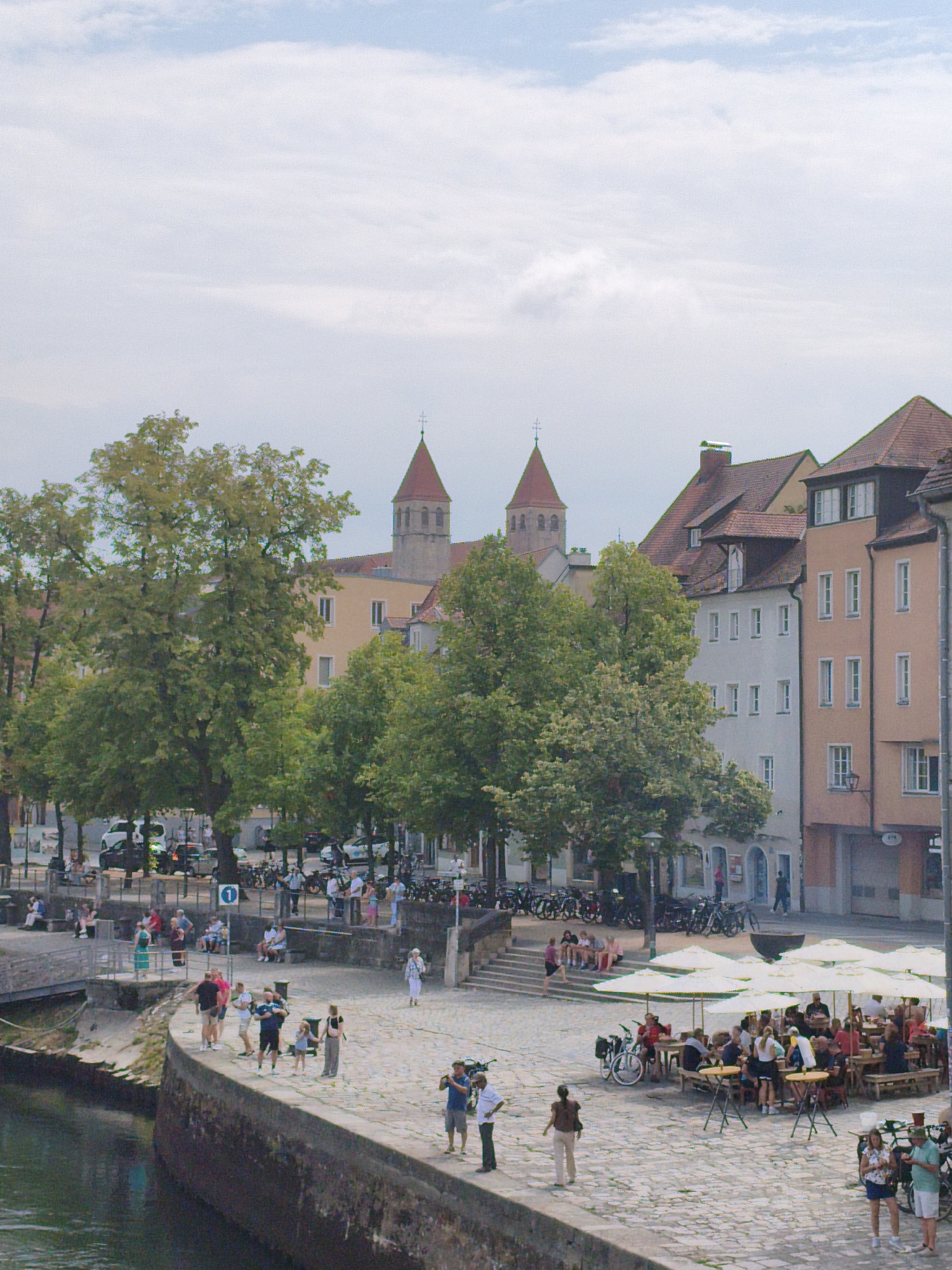 This image depicts a scene in the city of Regensburg, Germany, specifically along the Danube riverfront, likely near the Steinerne Brücke (Stone Bridge). The perspective is from across the river, looking towards the old town.
In the foreground, a stone embankment lines the dark water of the Danube. A paved cobblestone area extends from the embankment, populated by numerous people. Some individuals are standing and conversing, while others are seated at tables under large white umbrellas, indicating an outdoor cafe or restaurant area. Several bicycles are parked nearby.
Further back, large, mature trees with lush green foliage provide shade and are interspersed with buildings. The architecture suggests an older European city, with multi-story buildings featuring tiled roofs and various window styles. Two church towers with distinctive conical roofs are visible in the background, characteristic of Regensburg's historical churches, possibly St. Peter Cathedral or other churches in the Domberg area.
On the embankment, there are groups of people engaged in various activities: some are walking, others are sitting on benches or the edge of the embankment, and a few are standing near the water. The overall atmosphere is one of a bustling, pleasant summer day with outdoor leisure activities. The sky is partly cloudy, with a mix of blue sky and white clouds. The lighting suggests it is daytime, with soft sunlight filtering through the clouds.