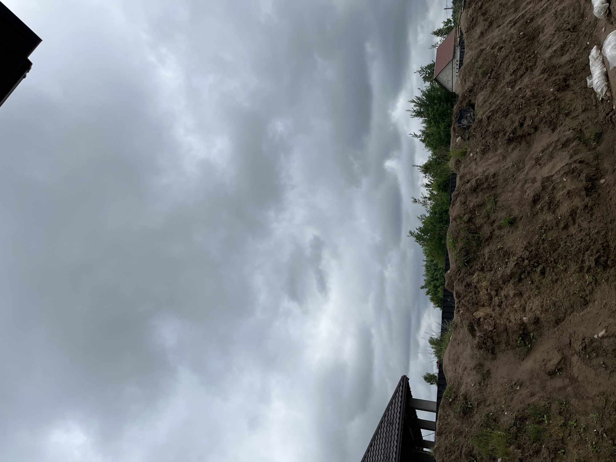 The image depicts a cloudy sky over a construction site. In the foreground, a large mound of earth, likely excavated during construction, dominates the lower portion of the frame. Scattered green vegetation, including grass and small plants, grows on the mound. In the middle ground, a row of trees forms a dense line. Beyond the trees, two houses are visible. The house on the left has a dark, tiled roof and is partially obscured. The house on the right has a red-tiled roof and appears to be constructed of light-colored materials. In the lower right corner, several white sacks are visible, possibly containing building materials or soil. The overall atmosphere is overcast and somber, suggesting the possibility of rain. The scene is situated in the Baravliany village council, Belarus.
