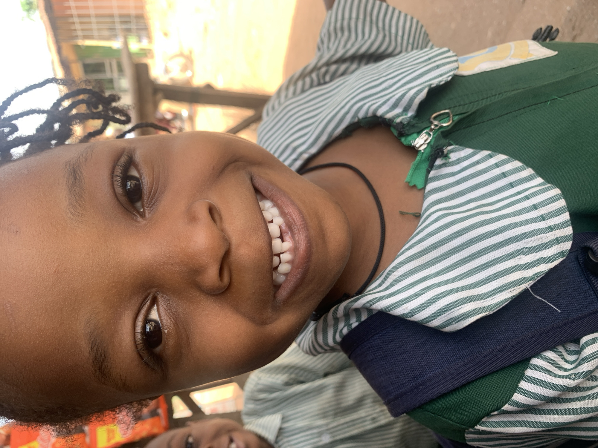A young, dark-skinned girl with braided hair is smiling broadly. She wears a green and white striped collared shirt, possibly a school uniform, with a dark blue underlayer. A black string necklace is visible on her neck. The photo is taken outdoors in what appears to be Sabon Tasha, Nigeria, based on the environmental cues (the building in the background), where similar uniform styles are common. A portion of another person's face is visible in the bottom center of the frame. The lighting is bright, suggesting a sunny day.