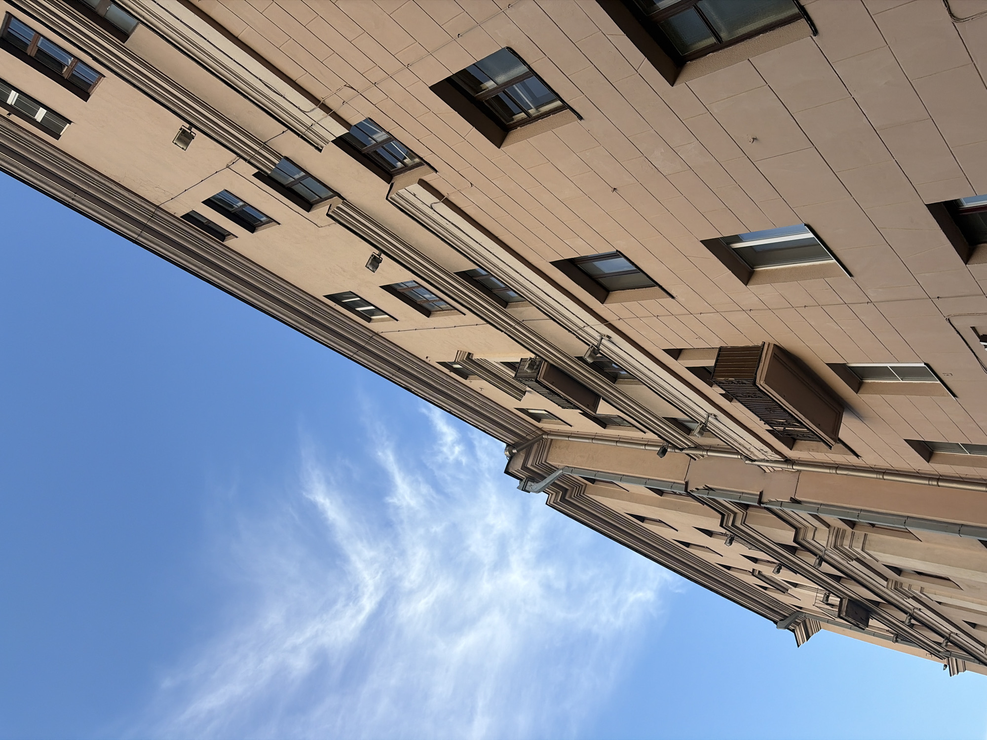 An upward-angled photograph shows a multi-story building against a bright blue sky with wispy white clouds. The building is constructed of beige stone blocks and features numerous windows framed in darker material. A balcony is visible on the right side of the building. Gutter pipes run vertically along the facade. The architecture has a classical, formal design. The image's perspective gives the impression that the building is leaning diagonally against the sky.