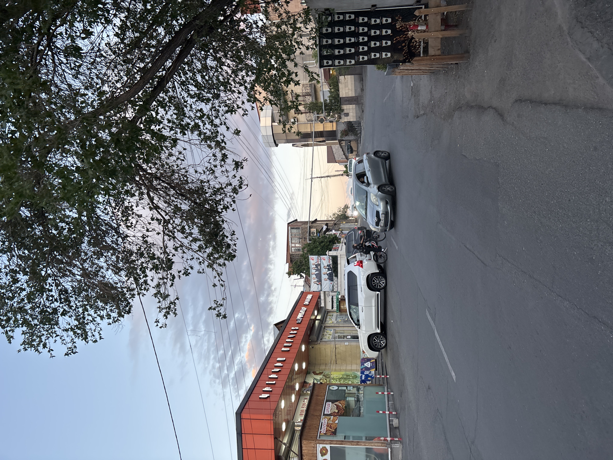 The image shows a street scene in Yerevan, Armenia, featuring several parked cars, a motorcycle, a storefront with Armenian signage, and a tree. The street is paved and has a white lane marker. Overhead power lines stretch across the sky. The storefront has a modern design with orange and white accents. The sky is partly cloudy with a hint of sunset colors. The overall setting appears to be a residential or commercial area within the city.