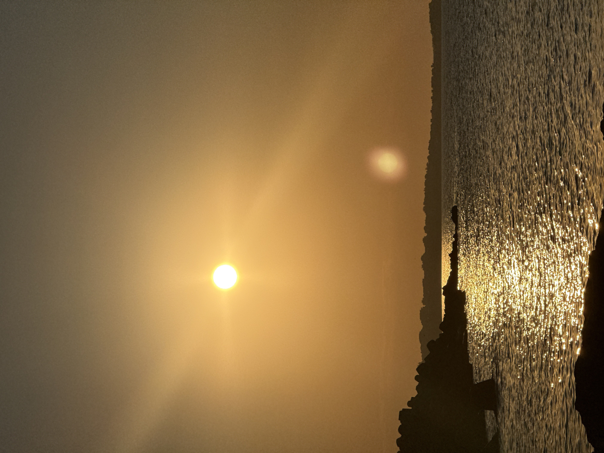 This is a vertical, outdoor, medium-shot photograph of the Black Sea coast at sunset, likely near Sozopol, Bulgaria. The sun, a bright orb of golden light, is positioned high in the orange-tinted sky, casting a shimmering path of light across the rippling water. In the foreground, dark, jagged rocks, possibly a breakwater or natural formations, line the left side of the frame, with the choppy water lapping against them. Silhouetted against the horizon is a faint, low-lying coastline with the suggestion of structures. The overall mood is serene and evokes the feeling of a tranquil evening by the sea. There are no people or animals visible in the image.