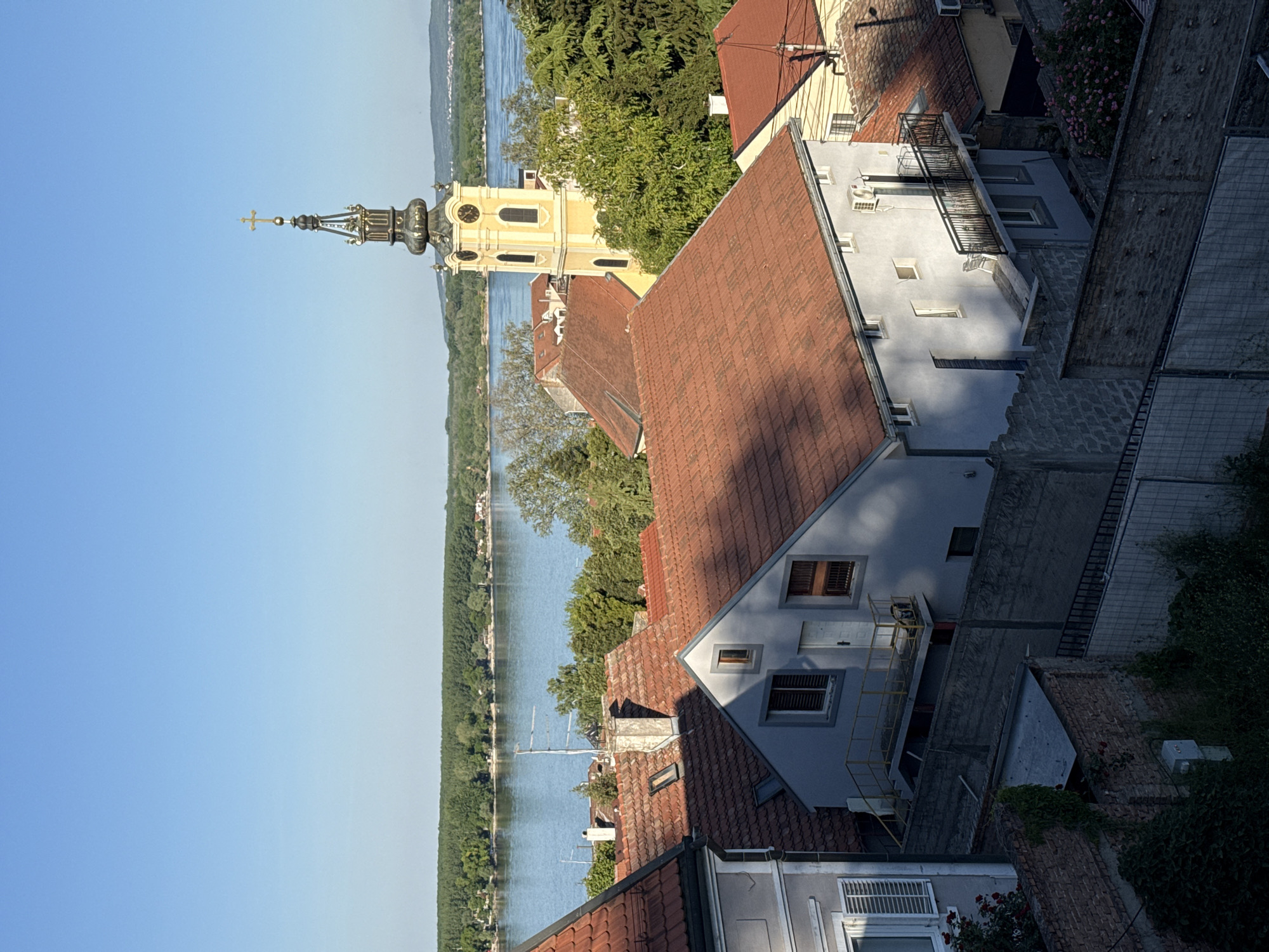 The image captures an elevated view in Zemun Urban Municipality, Serbia, overlooking a waterway lined with trees under a clear sky. Dominating the scene is a yellow church tower with a distinctive spire and clock face, rising above terracotta-tiled rooftops of adjacent buildings. These buildings vary in height and architectural style, with some featuring grey facades and balconies. The foreground showcases a mix of building materials, including brick and concrete. The overall impression is of a historic urban landscape nestled beside a riverbank.