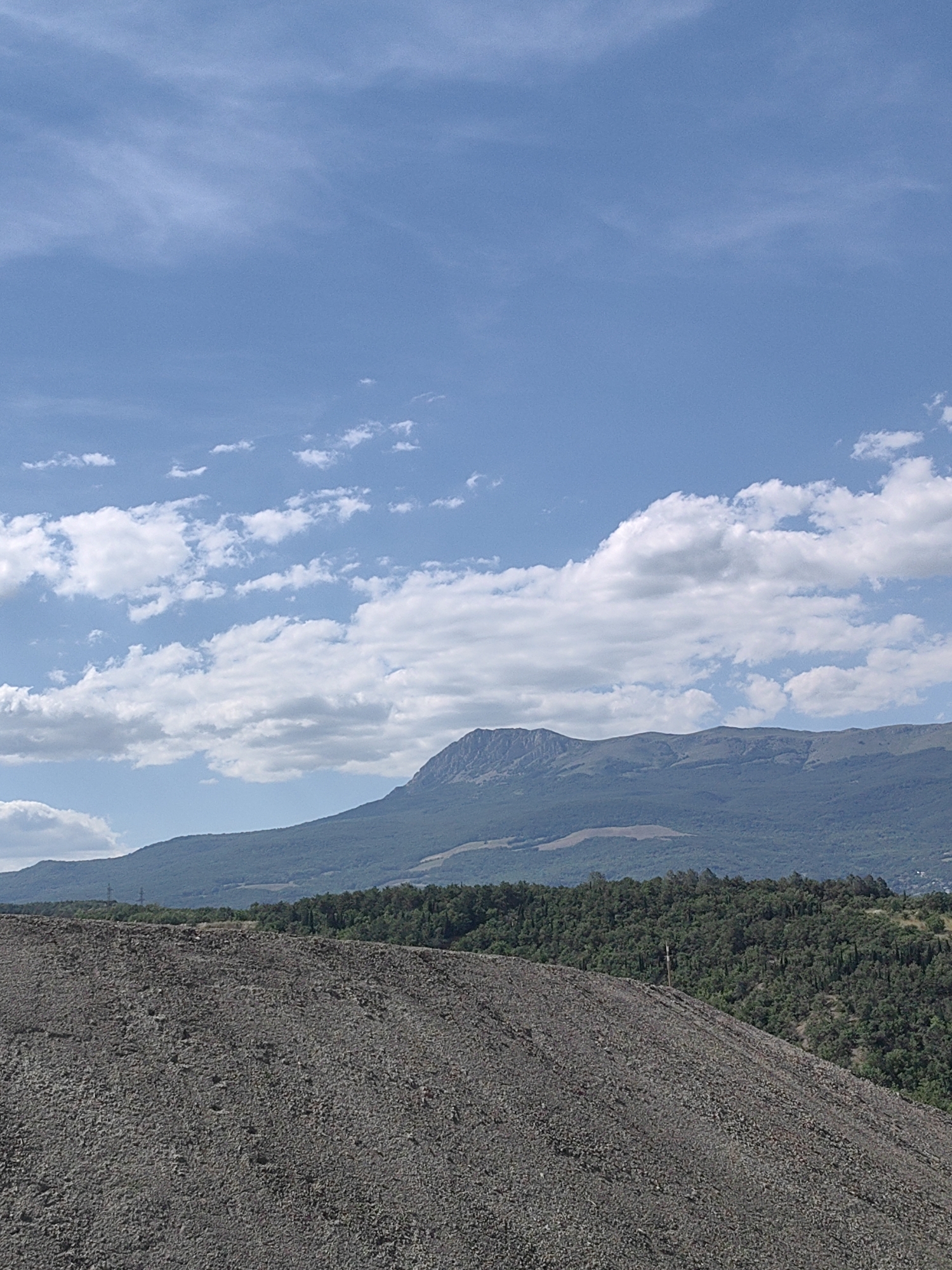 This media file depicts a landscape scene with a foreground of a large pile of dark-colored gravel or debris. Beyond the gravel pile, a dense, dark green forest covers rolling hills. In the background, a majestic mountain range stretches across the horizon, its peaks rugged and silhouetted against the sky. The sky is a vibrant blue, dotted with scattered white cumulus clouds that are illuminated by sunlight. The overall impression is one of a natural, somewhat wild environment. The lighting suggests it is daytime, likely late morning or early afternoon, with clear weather. There are no people or identifiable objects in the scene besides the natural elements and the gravel pile. The location appears to be rural or mountainous, consistent with the landscape cues. There is a small, light-colored cross or marker visible on the far right of the gravel pile, near the edge of the forest. There is also a faint, possibly man-made structure visible in the distance on the far left of the image, appearing as a vertical line against the distant hills. No text is visible in the image.