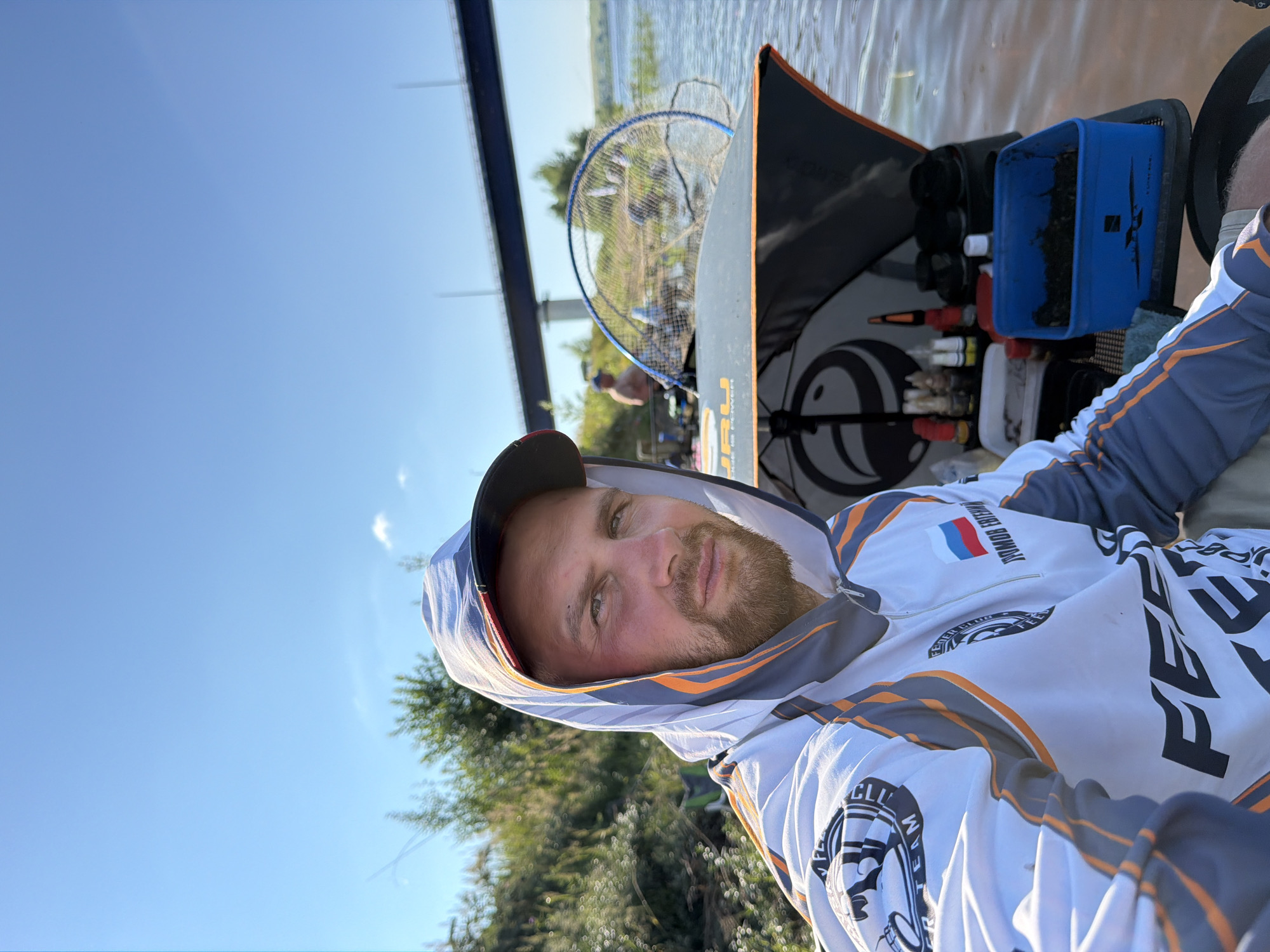 A man with a beard and a hooded jacket is sitting in a boat on a river. He is wearing a cap and looking at the camera. There are fishing supplies and a fishing net in the boat. In the background, there is a bridge and trees. The location is Tarasovo, Russia.