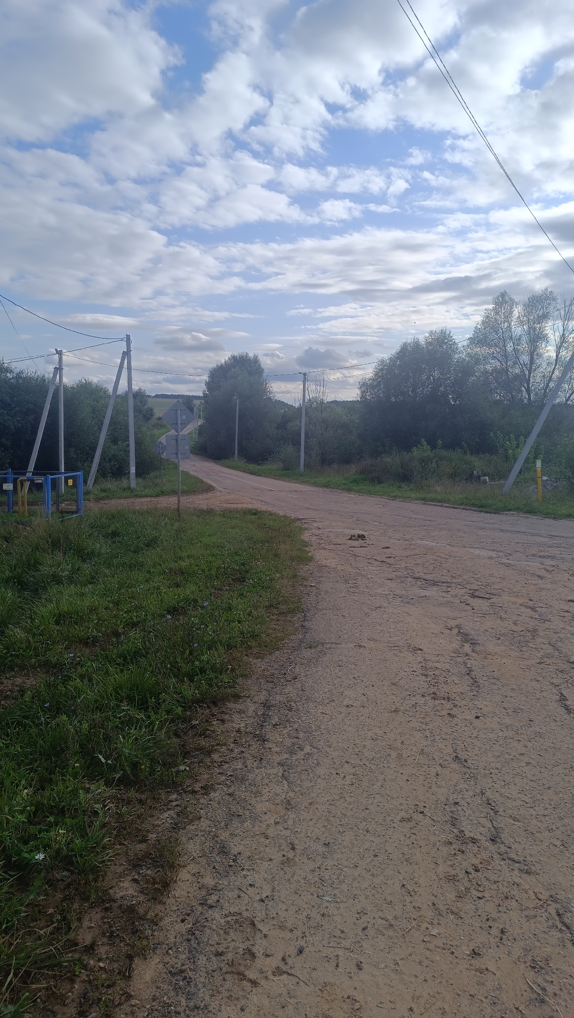 The image shows a dirt road curving to the right, bordered by green grass on the left and trees on both sides. In the distance, the road continues into a rural landscape with rolling hills under a partly cloudy sky. There are power lines overhead supported by wooden poles. A blue and yellow structure, possibly a gate or fence, is visible on the left side of the road near the foreground. A street sign is also present at the intersection. The overall scene depicts a rural road on a partly cloudy day.