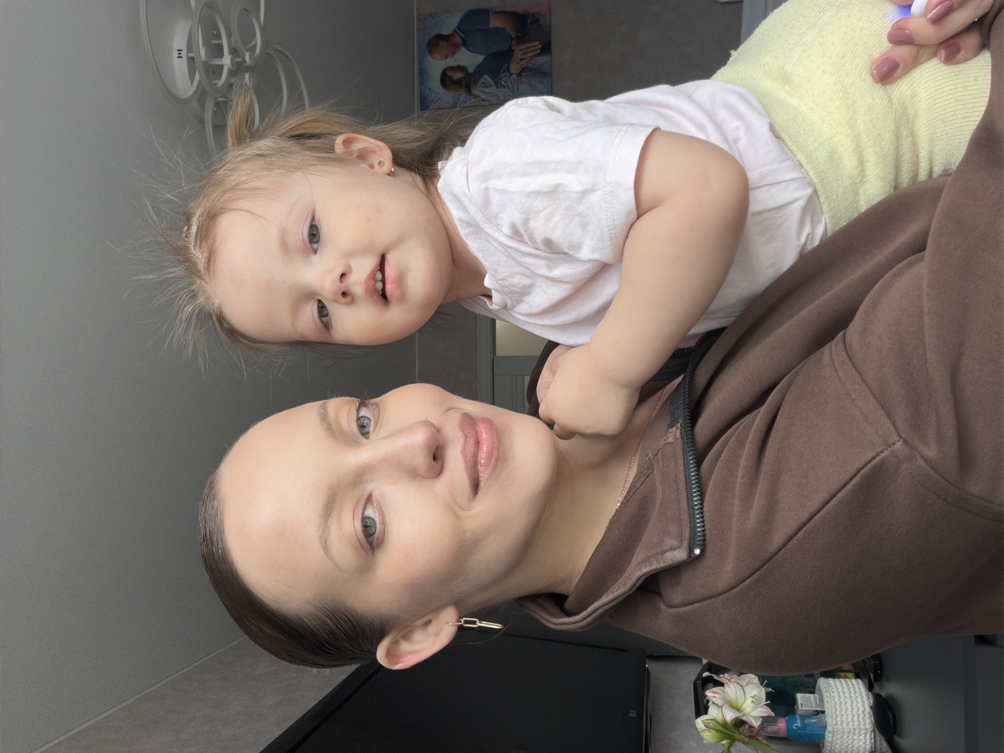 The image captures a close-up portrait of a woman and a young child in an indoor setting in Reshetnikovo, Russia. The woman, with light skin, light eyes, and dark hair pulled back, is wearing a brown zippered hoodie and a distinct gold-colored chain-link earring. She looks towards the camera with a subtle smile, her lips accented with light pink lipstick.

Perched or held above and slightly behind the woman's right shoulder is a young child, likely a toddler. The child has light hair tied in a small ponytail, light eyes, and is wearing a light pink t-shirt and light yellow bottoms, along with a small gold-colored stud earring. The child is also looking directly at the camera with a soft, engaging smile.

In the background, a plain grey wall is visible. A modern, circular light fixture is on the ceiling. On the wall behind the child, a framed picture shows two adults embracing. In the lower right corner, partially visible objects include a small bouquet of white and pink flowers, a blue bottle, and a white woven item, possibly a small basket or rolled towel, resting on a dark surface. The woman's hand, with pink manicured nails, is visible in the upper right, holding a small white object that emits a blue light.

The lighting is bright and even, indicating a daytime setting. Both subjects appear content and happy, conveying a warm, familial moment. No readable text is visible on any items in the image.