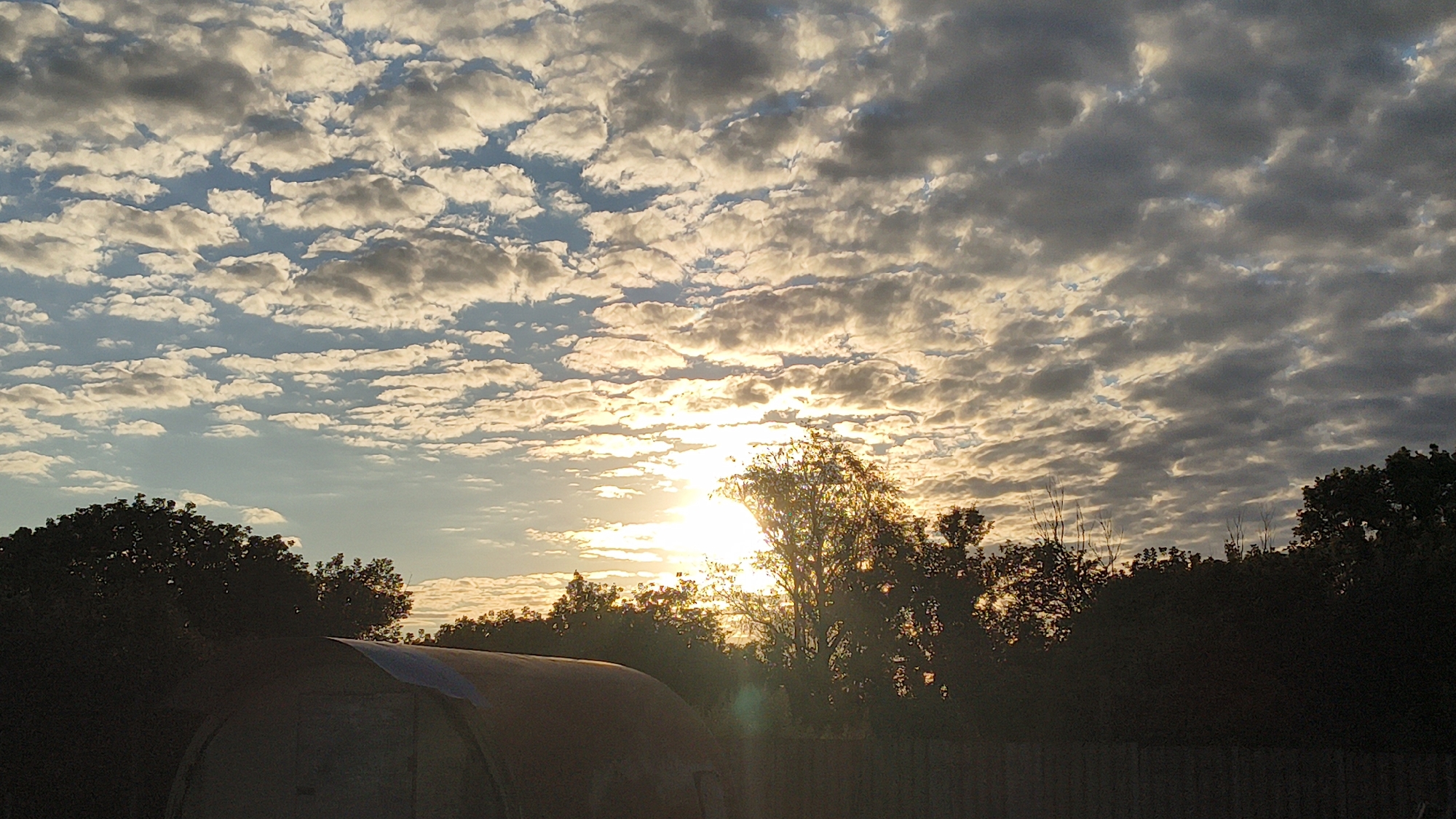 The image shows a sunset over a rural landscape in the city of Larinskii, Russia. The sky is filled with scattered clouds, with the sun breaking through in the center, casting a warm glow. Silhouetted trees line the horizon, and a dark, curved structure, possibly a greenhouse or a shelter, occupies the lower left portion of the frame. A wooden fence runs along the bottom edge of the image. The scene appears peaceful and natural, with no people or specific actions depicted.