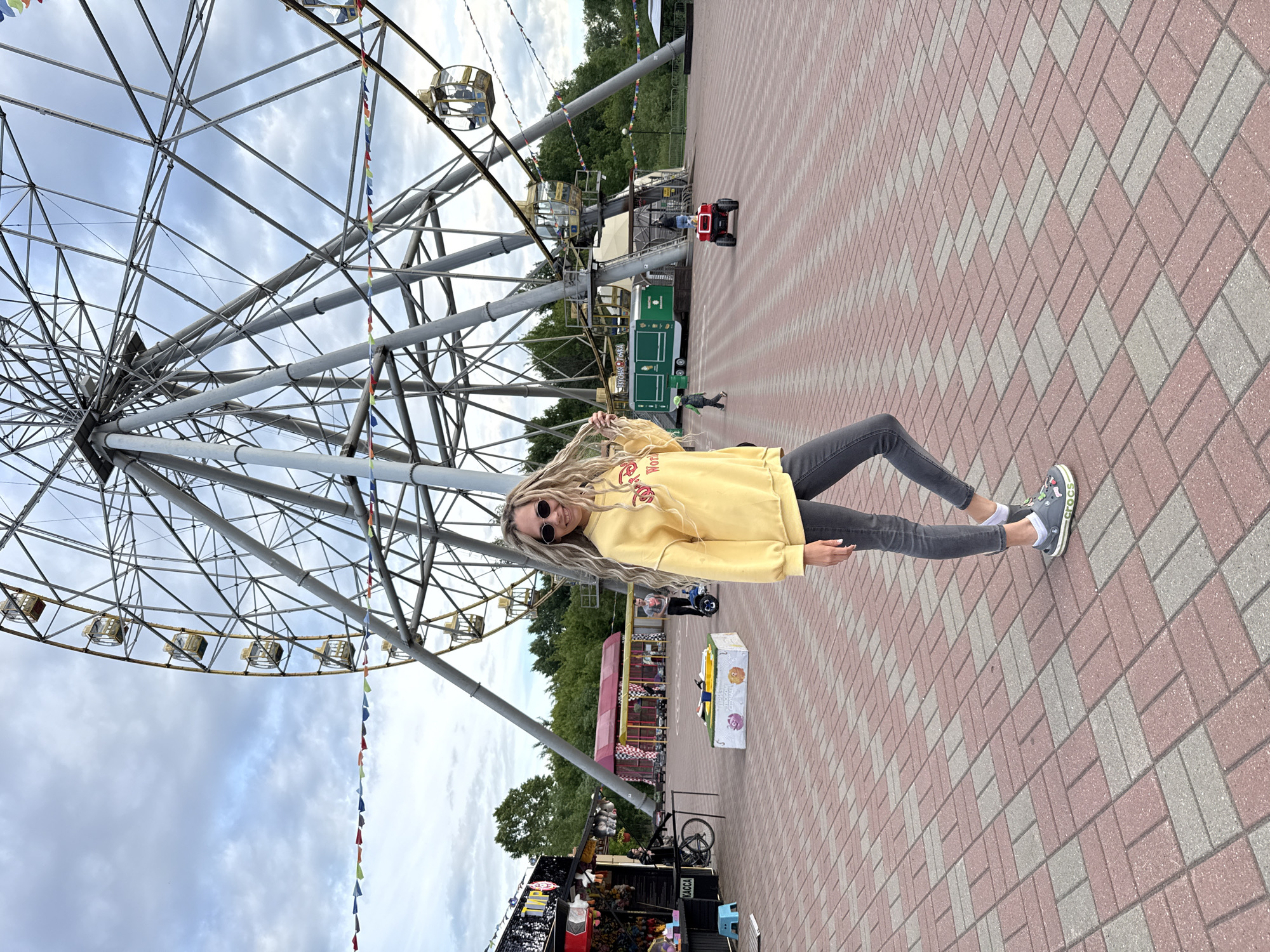 A woman with long blonde dreadlocks or braids, wearing dark sunglasses, a yellow long-sleeved sweatshirt featuring a red "Coca-Cola" logo, dark grey jeans, and grey Crocs with white socks, poses for the camera. She stands smiling, looking directly forward, on a paved ground made of alternating red and light grey rectangular bricks laid in a diagonal pattern.
Behind her, a large metal Ferris wheel dominates the left side of the frame, its intricate spokes and several empty gondolas visible against an overcast sky with scattered clouds. Colorful triangular bunting hangs from one of the Ferris wheel's supports.
The scene depicts an amusement park or carnival. In the mid-ground, several attractions and stalls are present. To the right of the woman, a green food truck or stall has the word "SUGAR" partially visible on its side. Further back, another stall displays "KACCA" (Касса), indicating a cashier or ticket booth in Russian. Green trees are visible in the background, along with other amusement park structures and a small red toy car on the pavement. The setting suggests a casual, cheerful daytime outing. The location is Шебунино, Russia.