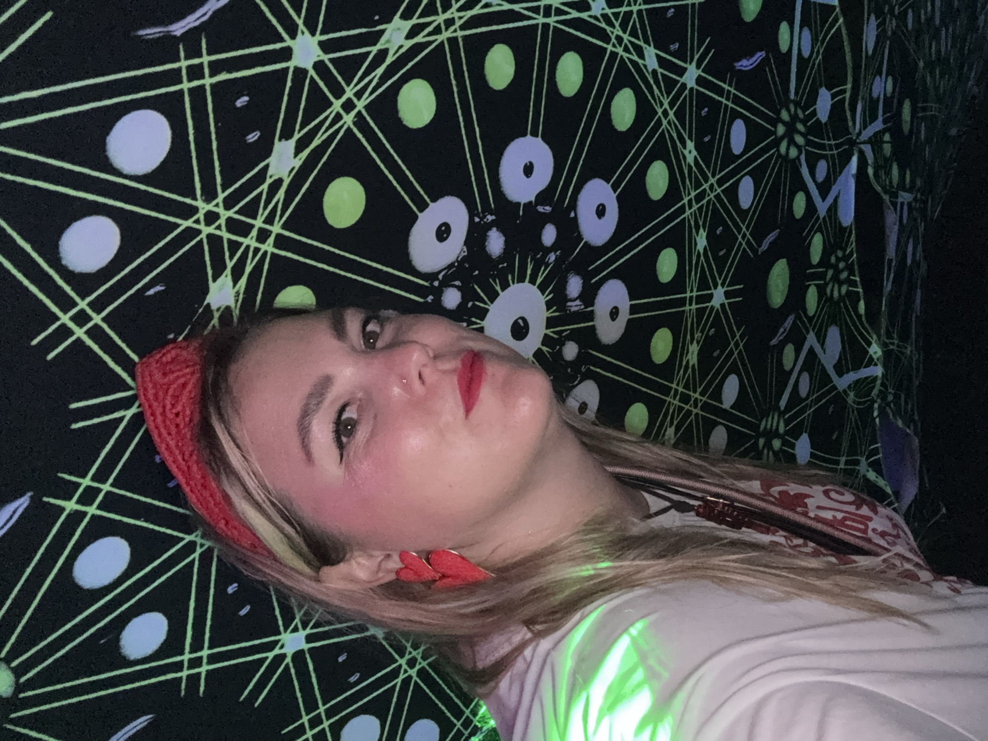 This is a selfie taken by a blonde woman with red heart-shaped earrings and a red headband, wearing a white t-shirt. She is standing in front of a black backdrop with a neon green geometric pattern and scattered circles of light blue and green. The pattern resembles a starburst or a spider web. The lighting is dim with green light cast on the woman's shoulder and the background. The location is not specified in the image or the provided context.