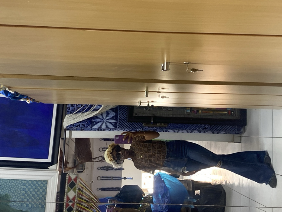 An individual is depicted taking a mirror selfie indoors. The subject, appearing to be female, is positioned centrally in the reflection, wearing sunglasses, a light-colored curly hat or wig, a short-sleeved brown top with a repeating geometric pattern, dark blue ripped flare jeans, and dark shoes. A golden necklace with a square pendant is visible. The subject holds a purple smartphone in their right hand, angled towards the mirror.
The reflected background reveals a diverse interior. To the reflected subject's left, a voluminous blue object, possibly fabric or a bag, rests on a dark surface. Behind the subject, multiple dark-colored items, possibly bags or keychains, are suspended against a light-colored wall. Above, various framed artworks or textile pieces are displayed, including a prominent blue abstract painting and other patterned designs. A section of the wall to the reflected subject's right features blue and white patterned tiling or fabric with a starburst motif. The floor is light-colored and reflective.
The right side of the image, not part of the reflection, shows a series of vertically oriented light-colored wooden cabinets or lockers. Each cabinet features a chrome handle with a key inserted into its lock. A blue textile item is partially visible atop the cabinets in the upper right corner.
The setting suggests a commercial space, such as a boutique, showroom, or a retail store, possibly incorporating a changing room or storage area. The specific location in the city of Alaguntan, Nigeria, cannot be confirmed from visual evidence within the image.